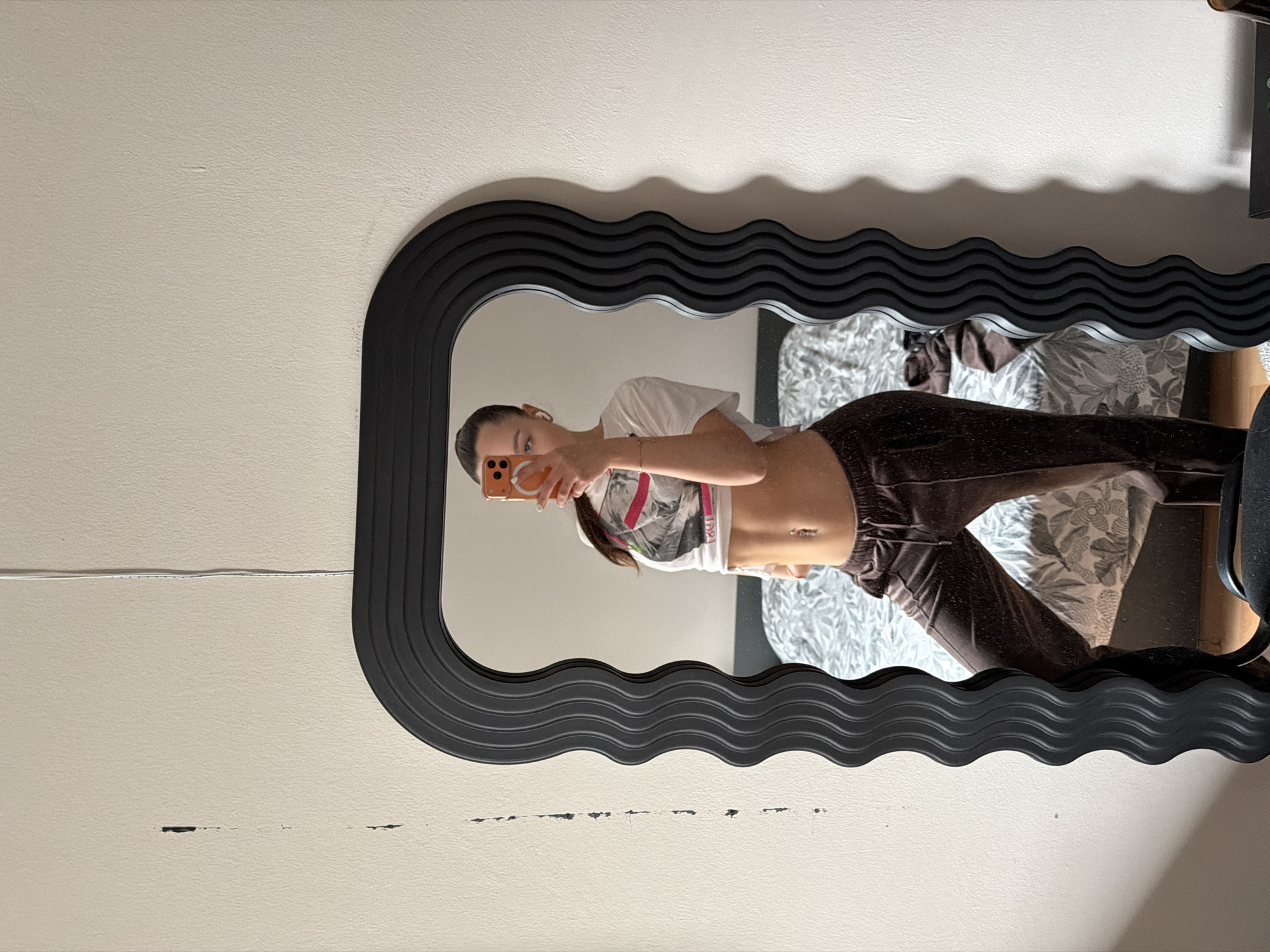 A young woman is depicted taking a mirror selfie in an indoor setting, identified as Sofia, Bulgaria. The image, originally rotated, shows her reflection in a large, vertically oriented mirror with a distinctive black, wavy, and rippled frame.
The woman, with dark hair pulled back and light eyes, holds an orange-cased smartphone in her right hand to capture the image. She is wearing a white, short-sleeved crop top featuring a graphic design with some indistinct text and numbers, possibly "1991" or "1971". Her midriff is visible, adorned with a belly button piercing. She pairs the top with dark brown, possibly velvet or corduroy, pants.
The background reflected in the mirror reveals a bed with a patterned duvet or blanket, indicating a bedroom setting. A dark piece of furniture, possibly a chair, is also visible to the right within the reflection. The mirror is mounted on a plain, light-colored wall with a textured finish. A thin white cable runs horizontally across the wall above the mirror, and several small scuffs and marks are visible on the wall, particularly below the mirror frame. The lighting suggests an interior environment, likely illuminated by artificial light.