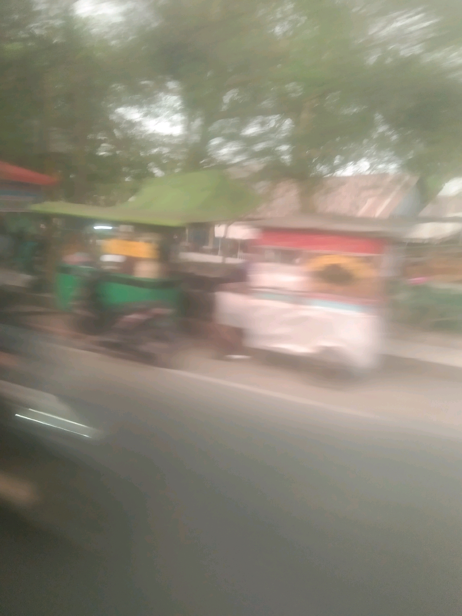 The image shows a street scene in Tanjung Raja, Indonesia. The photo is blurry, suggesting it was taken from a moving vehicle. In the foreground, a gray road surface is visible, with some streaks of light indicating movement. Behind the road, there are several roadside stalls or small shops. These structures have green and red awnings, and some appear to be selling goods, with yellow and white objects visible within them. Trees with green foliage are seen in the background, and the sky is overcast. There are no clearly identifiable people or specific landmarks that pinpoint the exact location within Tanjung Raja, but the overall impression is of a typical street scene in a busy urban or semi-urban area of Indonesia.