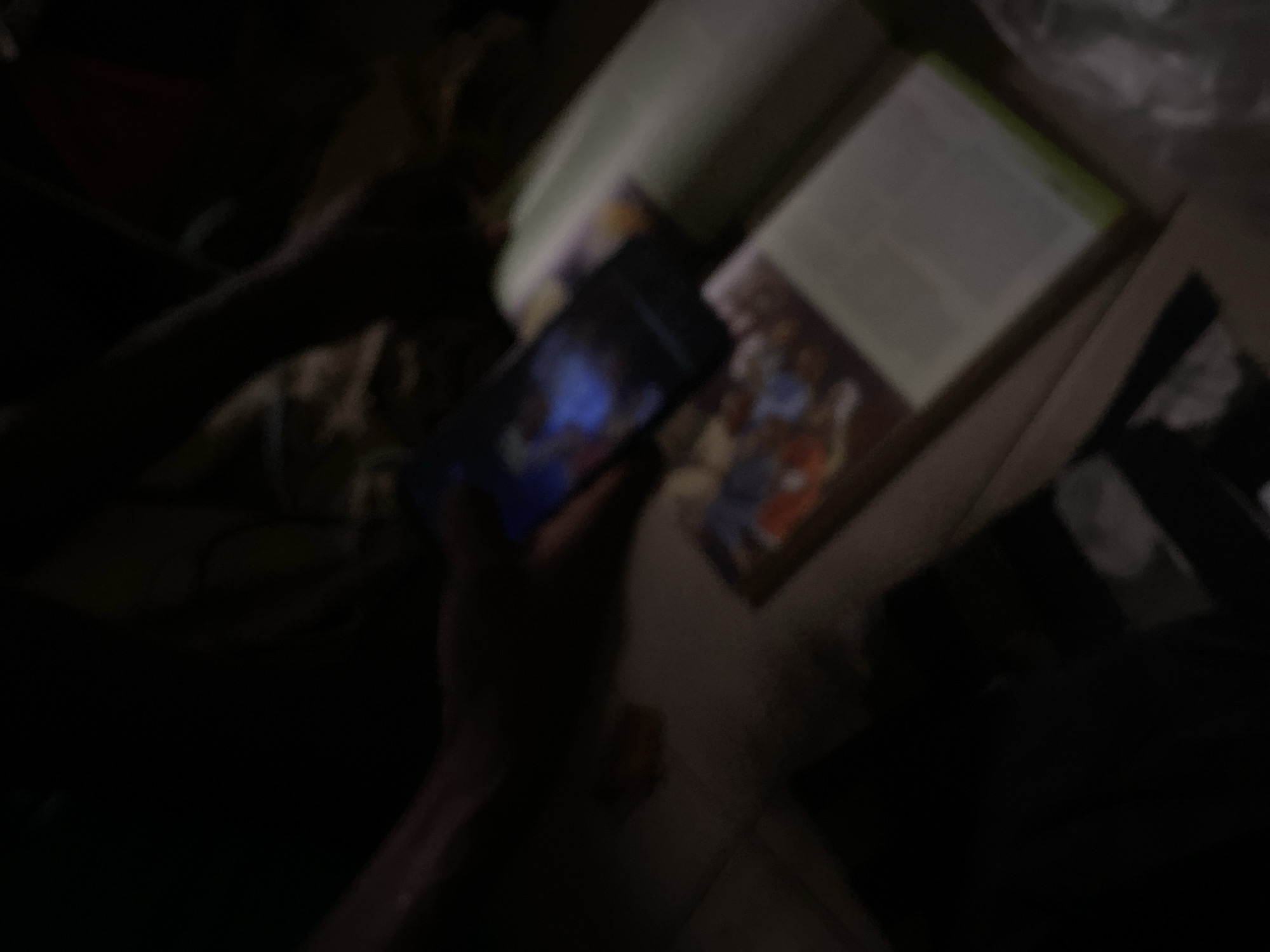A dimly lit, blurry image captures a person's hands holding a smartphone displaying a vibrant image, possibly a digital artwork or game screen with blue and purple hues. Below the phone, an open book with illustrated pages is visible. The book's pages are mostly white with some colorful illustrations. The overall scene suggests a relaxed indoor setting, possibly a home or a quiet study area. The image lacks specific details to pinpoint a precise location within Jauro Yinu, Nigeria, but it depicts a moment of personal engagement with digital media and reading.