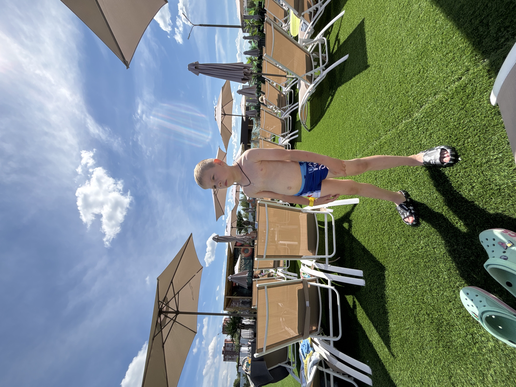A young boy stands on artificial turf in a resort or water park area. He is wearing blue swim trunks and a yellow wristband. In the background, there are rows of empty lounge chairs and several umbrellas, suggesting a day at a pool or beach. The sky is blue with scattered white clouds. The scene appears to be outdoors on a sunny day.