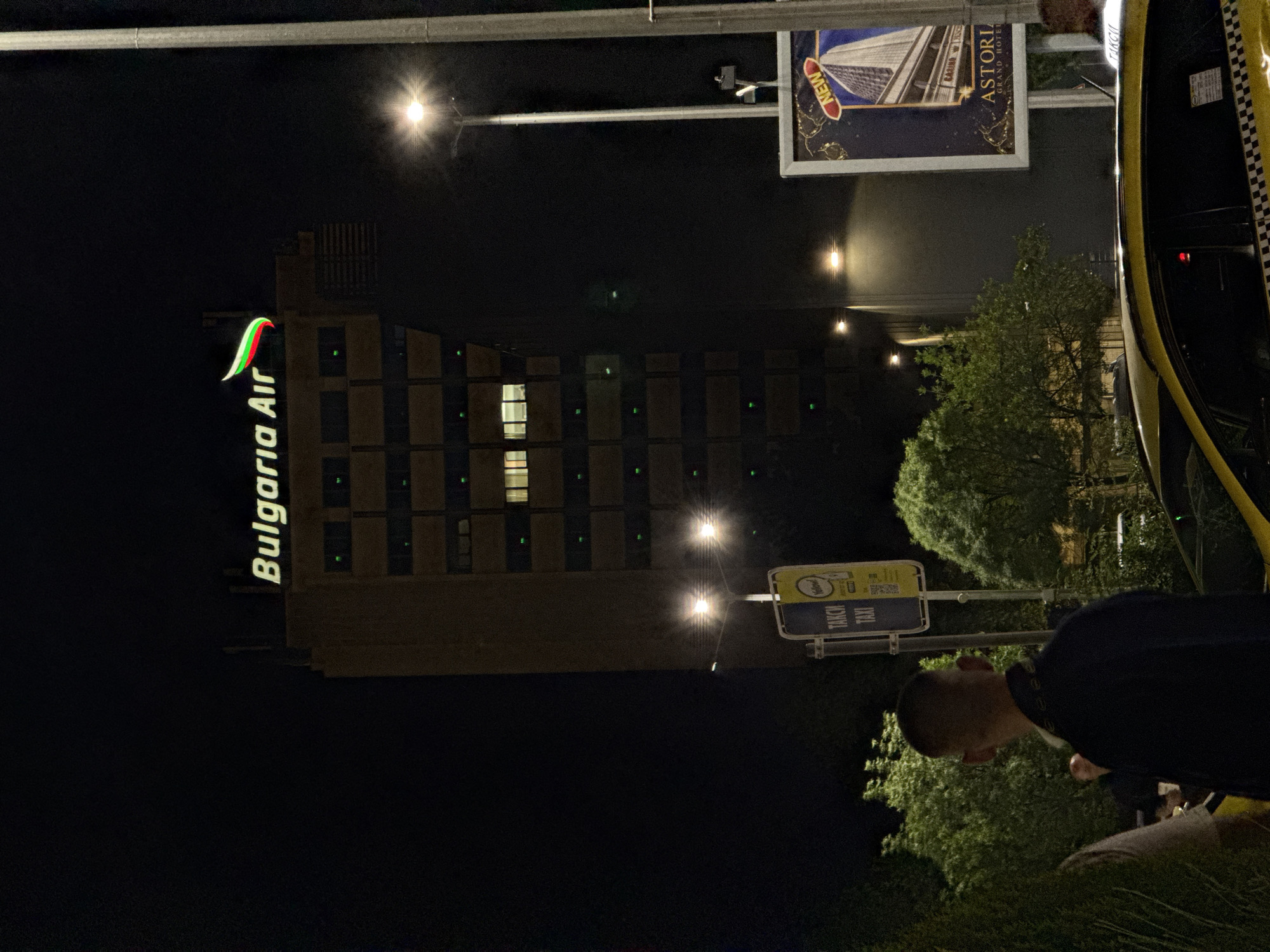 The image captures an urban night scene, likely on a street or public square in Sofia, Bulgaria. The primary subjects include a large, multi-story building, street furniture, a taxi, and partially visible individuals.
The imposing building, likely a hotel or commercial structure, stands on the left, illuminated by scattered internal lights, with one window shining brightly. Vertically mounted on its facade is a prominent, illuminated sign displaying "Bulgaria Air" alongside a distinctive green, white, and red stylized logo, reminiscent of the Bulgarian flag.
On the right side of the frame, the yellow and black checkered roof of a taxi cab is visible, indicating its presence at what appears to be a taxi stand. Nearby, a blue and yellow sign clearly displays "TAKCИ" (Taxi in Cyrillic) multiple times, confirming the taxi service location. Further right and above, an illuminated billboard-style sign advertises "ASTORIA GRAND HOTEL" with the word "NEW" also visible, displaying an architectural image.
Several bright streetlights or overhead lamps cast stark illumination, creating star-like flares across the dark sky. Green foliage from trees is visible towards the right and bottom right, adding a natural element to the urban setting. A person with short hair, wearing a dark top, is partially visible in the bottom right, facing downwards or away from the camera. Another individual's head is barely discernible in the top right corner.
The scene captures a moment of nighttime activity, characterized by artificial lighting, commercial signage, and the presence of transportation services in a city environment.