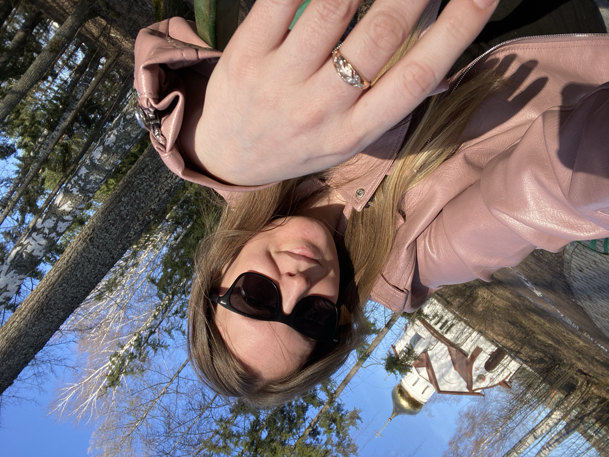 A woman wearing sunglasses and a pink leather jacket is in an outdoor setting, likely a park or area with trees. A white building with a golden dome, possibly a church or cathedral, is visible in the background. The picture may have been taken in Noviy, Russia. The woman's hand with a ring is in the frame.