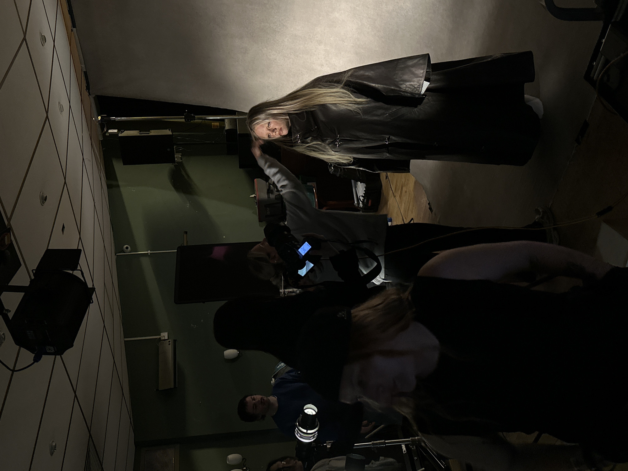 This image depicts a dimly lit indoor studio setting, possibly a photography or film set, in London, United Kingdom. The primary subject is a young woman with long, blonde hair, wearing a dark, long coat. She is standing on a wooden floor in front of a large, light-colored backdrop.
Several other individuals are present, some appearing to be part of the production crew. One person, wearing a dark sleeveless top and a dark hat, is partially visible in the foreground, with their back to the camera. Another individual, wearing a grey hoodie, is seen with their hand near the blonde woman's head, potentially adjusting her hair or a piece of clothing. To the left, a person in a blue sweater is visible, facing towards the center of the scene. There are also various pieces of film and photography equipment, including lights and stands, scattered around the room.
The environment suggests a professional setup for capturing images or video. The lighting is dramatic, with strong spotlights illuminating the main subject and creating shadows elsewhere. The overall atmosphere is one of creative production.
