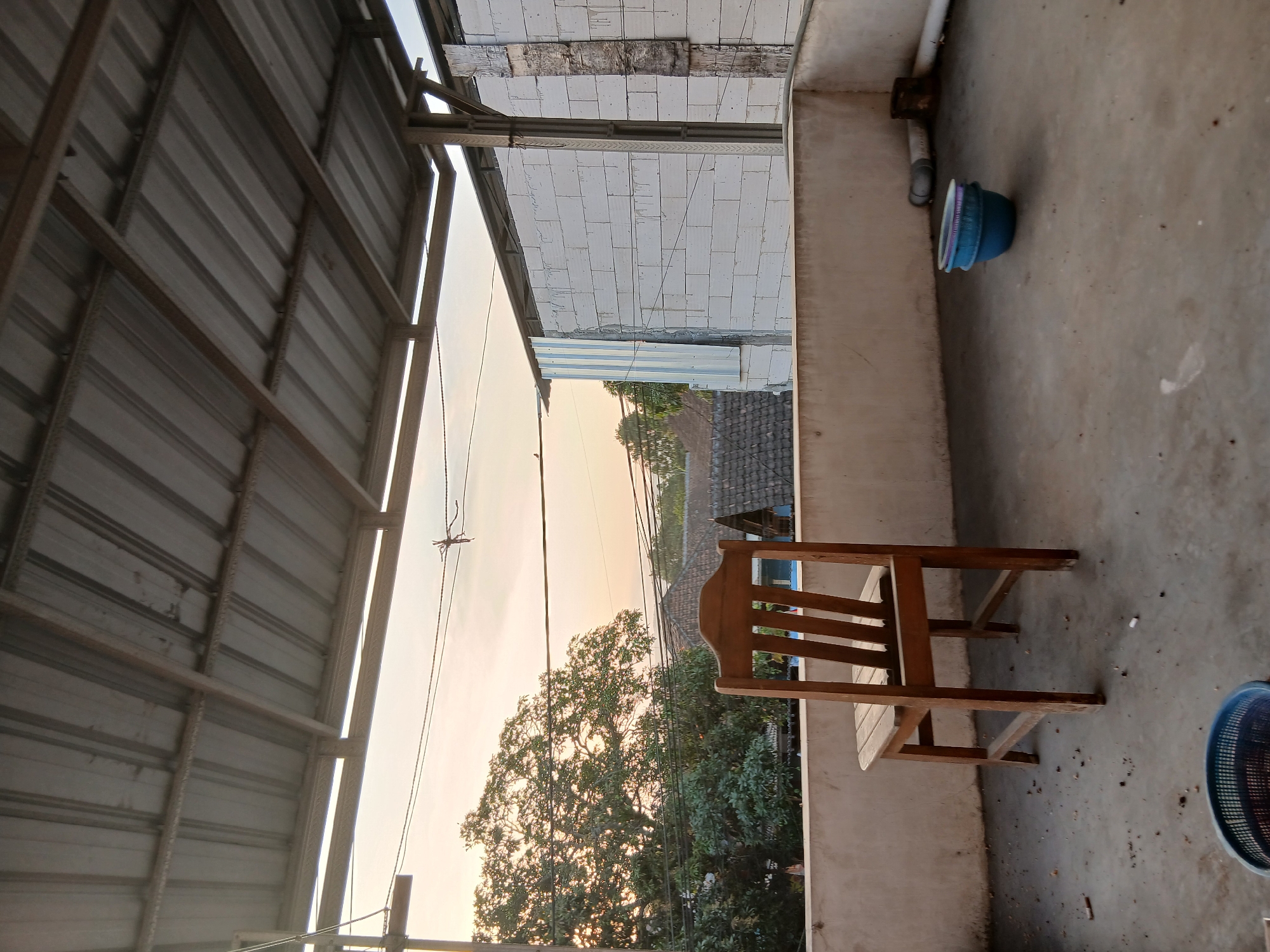 The image depicts an outdoor concrete patio area, likely a rooftop or balcony, in Tenggarang, Indonesia. A wooden chair stands prominently in the foreground. The patio is partially covered by a corrugated metal roof supported by metal beams. In the background, a large tree with dense foliage is visible, along with rooftops of houses, suggesting a residential area. The sky exhibits the soft hues of either sunrise or sunset. The patio floor is dusty and scattered with cigarette butts, indicating potential outdoor smoking. On the right side of the patio, there are some PVC pipes and a blue plastic basin. The wall behind the patio appears to be constructed from white cinder blocks. The scene captures a moment of stillness under a twilight sky.