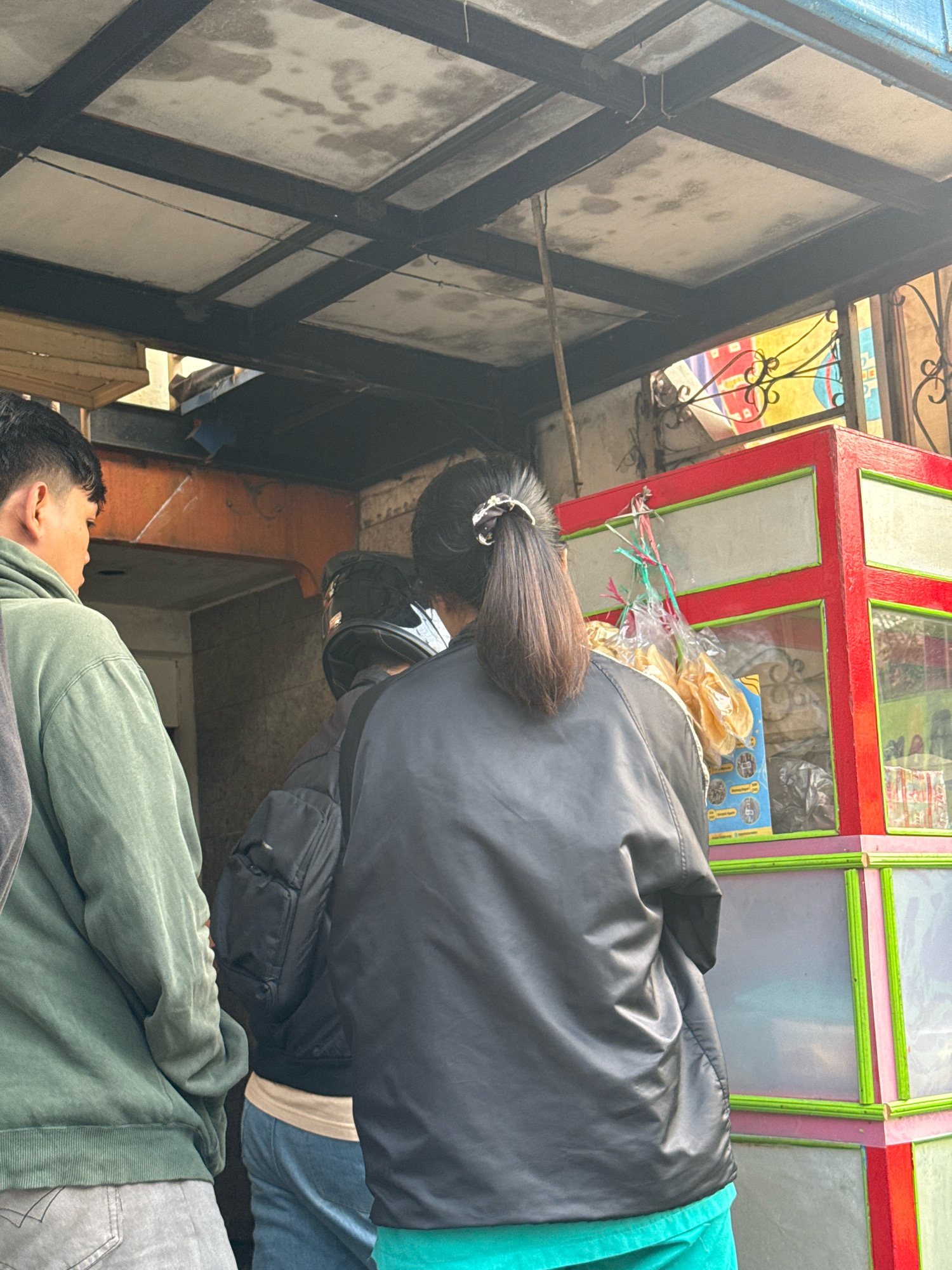The image depicts a street scene in Surabaya, Indonesia, focusing on a group of people near a food stall. In the foreground, a person wearing a black jacket faces away from the camera, with their dark hair tied in a ponytail with a white scrunchie. To their left, another individual in a green hooded sweatshirt is partially visible from the side. Behind the person in the black jacket, someone wearing a black helmet stands, facing the food stall. The food stall itself is a colorful structure with red and green accents and glass panels displaying various food items, including bags of snacks. The environment suggests an outdoor setting, possibly a busy market or street corner, with a ceiling structure visible above the stall.