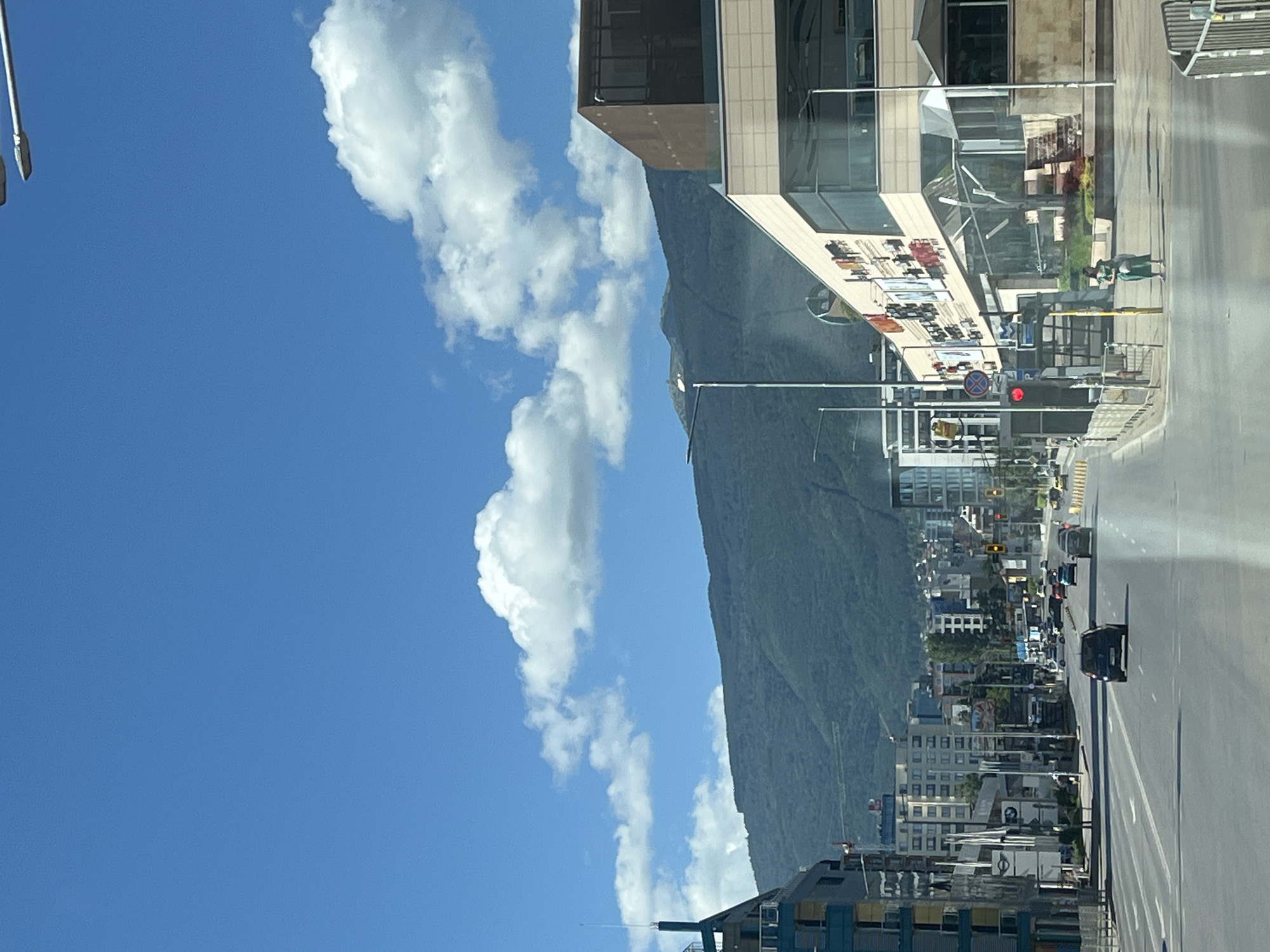 The image shows a wide, multi-lane road in a city. The road is lined with buildings on both sides, ranging from modern commercial structures with glass facades to taller residential buildings. A large, tree-covered mountain range dominates the background, with a clear blue sky dotted with fluffy white clouds overhead. Several cars are driving on the road, and a few pedestrians are visible on the sidewalks. Traffic lights and street lamps are also present. The scene appears to be a typical urban environment during daylight hours.