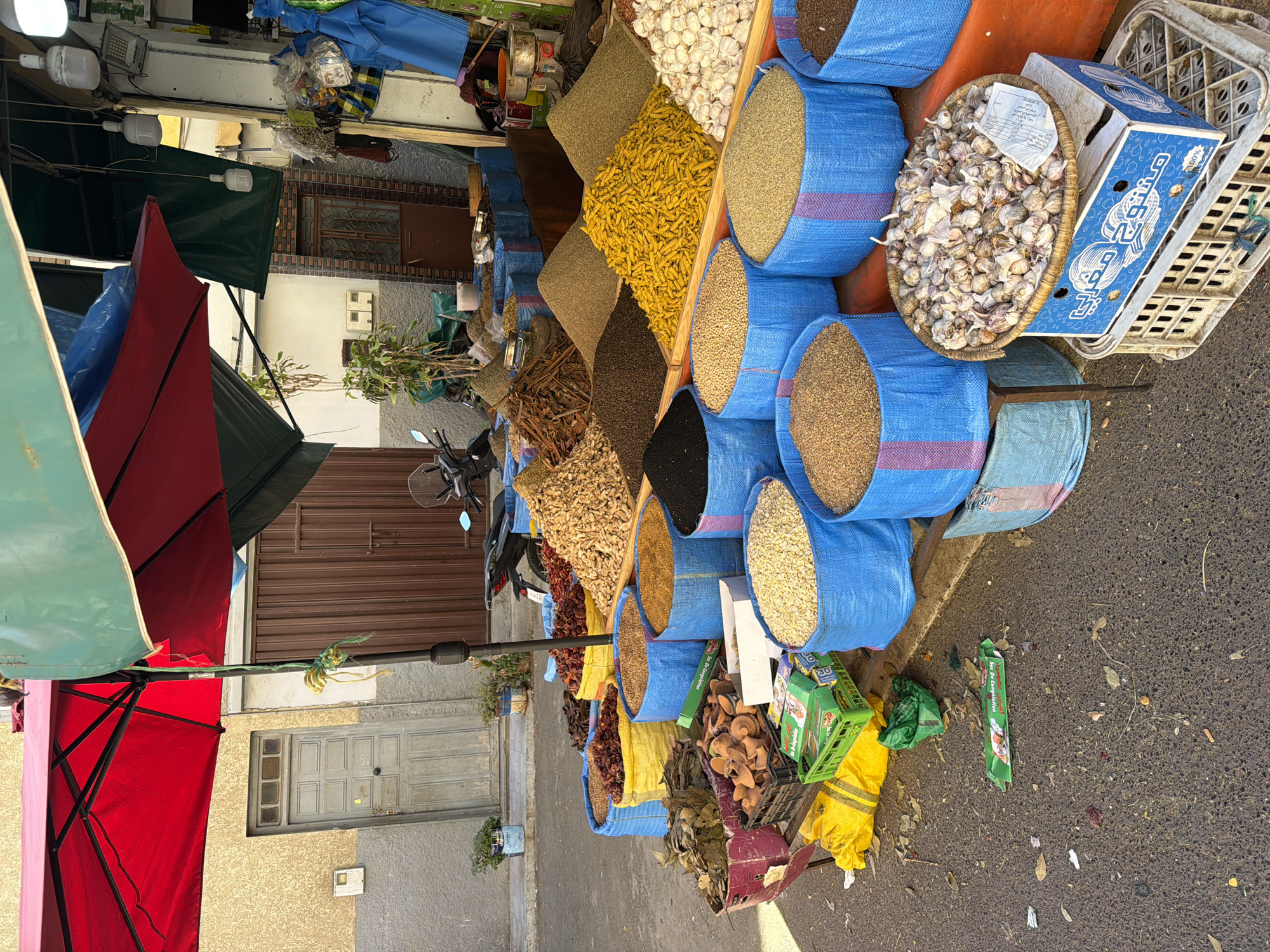 The image shows a spice market stall on a street in Kenitra, Morocco. Blue sacks filled with spices such as turmeric, coriander seeds, and black peppercorns are displayed. Garlic cloves in a wicker basket, and a crate with Arabic writing on it are also visible. The stall is shaded by a red and green umbrella. A motorcycle is parked nearby, and doors to buildings are in the background. Litter is scattered on the ground.