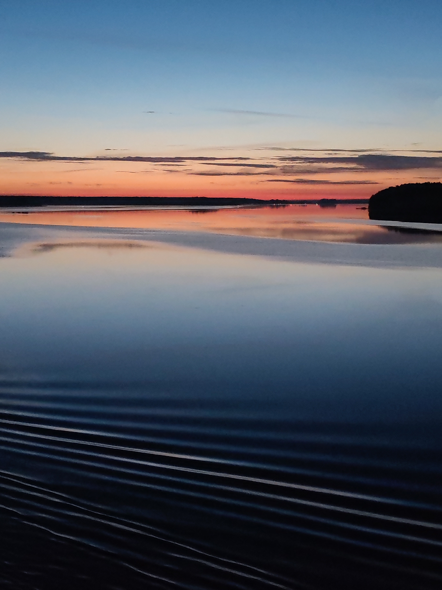 The image presents a still body of water, likely a lake or wide river, at twilight or dawn. The sky transitions from a deep blue at the top to hues of orange and pink near the horizon, where thin clouds are visible. The water reflects the colors of the sky, creating a mirror effect. In the distance, a dark treeline or landmass is discernible on the right side of the frame. The lower portion of the image exhibits regular, linear wave patterns, likely the wake of a moving vessel. Given the location is Nova, Russia, it could be a lake or river in the region.