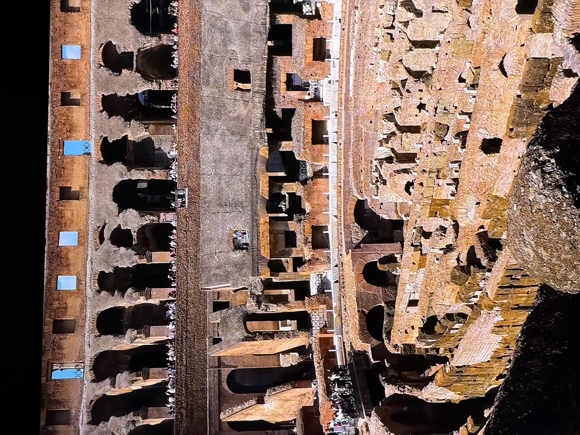 The image shows an interior view of the Colosseum in Rome, Italy. The structure is made of weathered brick and stone, with multiple levels of arches and decaying walls. In the foreground, there are tourists gathered on elevated platforms, observing the ruins. The lighting suggests it is daytime, with shadows cast by the structure. The environment is ancient and historical. The city of Chirchik, Uzbekistan, is not depicted in this image.