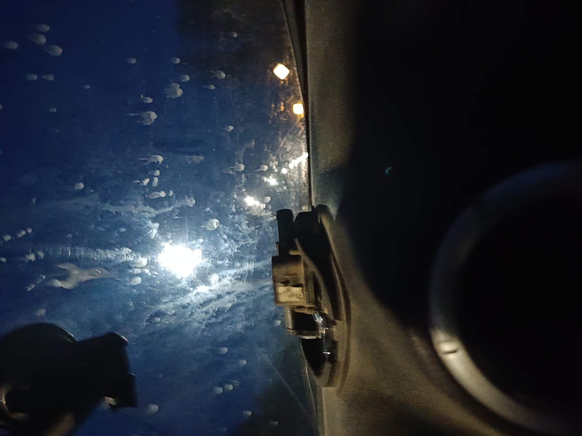 The image captures a nighttime scene from inside a vehicle in Chaikovsky, Russia. The left two-thirds of the frame show a dark blue exterior surface, likely a windshield or window, heavily covered with numerous irregular white droplets and streaks, consistent with water or foam residue. Through this partially obscured surface, several artificial light sources are visible: a prominent, circular bright white light in the lower-middle-left, and two distinct rectangular yellow lights positioned higher and further right. Additional smaller, fainter white lights are scattered across the blue surface. The right third of the image shows the vehicle's dark interior, including a light brown or tan mechanical component with visible intricate details, mounted near the center-right. Further to the right, a dark, curved element of the vehicle's interior is present. A dark, indistinct silhouette, potentially an object or a hand, occupies the lower-left foreground. No individuals are explicitly visible.
