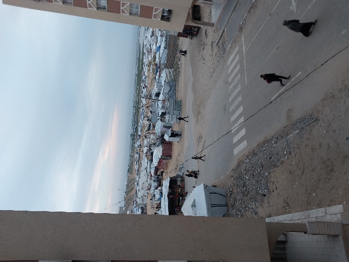 An elevated view shows a large camp of makeshift shelters extending to the horizon. A road with pedestrian crossings runs along the edge of the camp. Several people walk along the road and through the camp. Tall buildings overlook the scene. The sky is overcast.