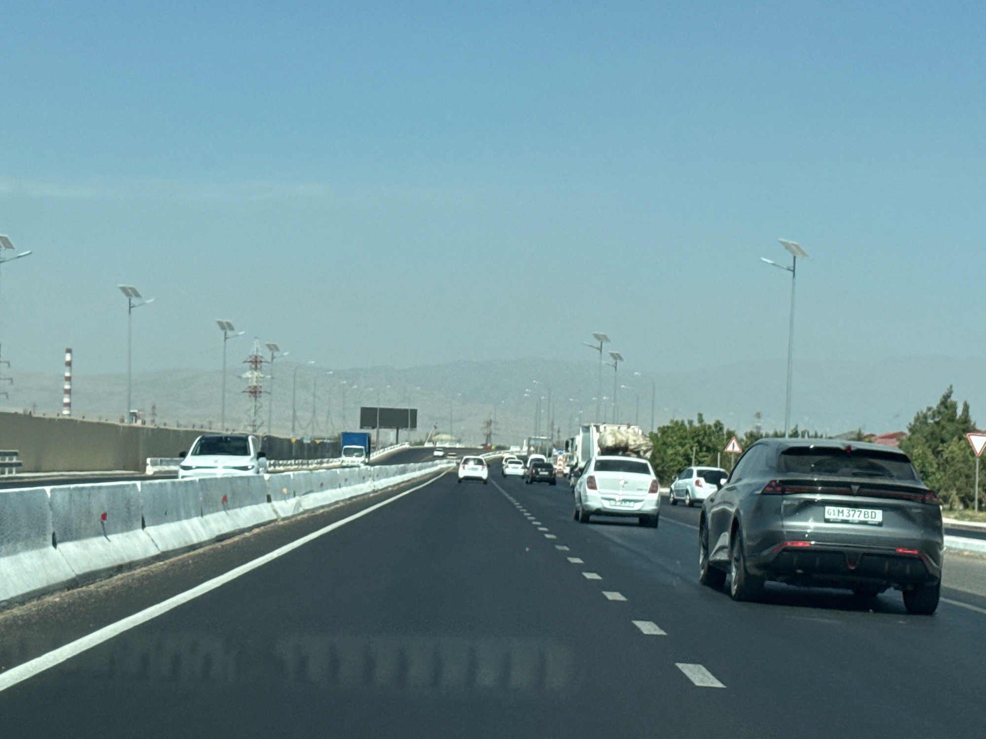 The image captures a view from a vehicle traveling on a multi-lane highway on a clear day. The road surface is asphalt, marked with white dashed lines. On the left side of the road, a concrete barrier separates the highway from an adjacent area with construction materials, industrial structures, and what appear to be solar-powered streetlights. The landscape in the distance is characterized by rolling hills and a hazy sky. Several vehicles are visible on the highway, including a white car, a blue truck, and other passenger vehicles, some of which are in the process of overtaking. In the foreground, a dark gray SUV is prominently featured, displaying a license plate that reads "G1M 377BD". The overall scene suggests urban or semi-urban development in a dry, possibly arid region. The presence of the specific license plate format points to the location being in Uzbekistan, potentially near Jaukashty, given the context.