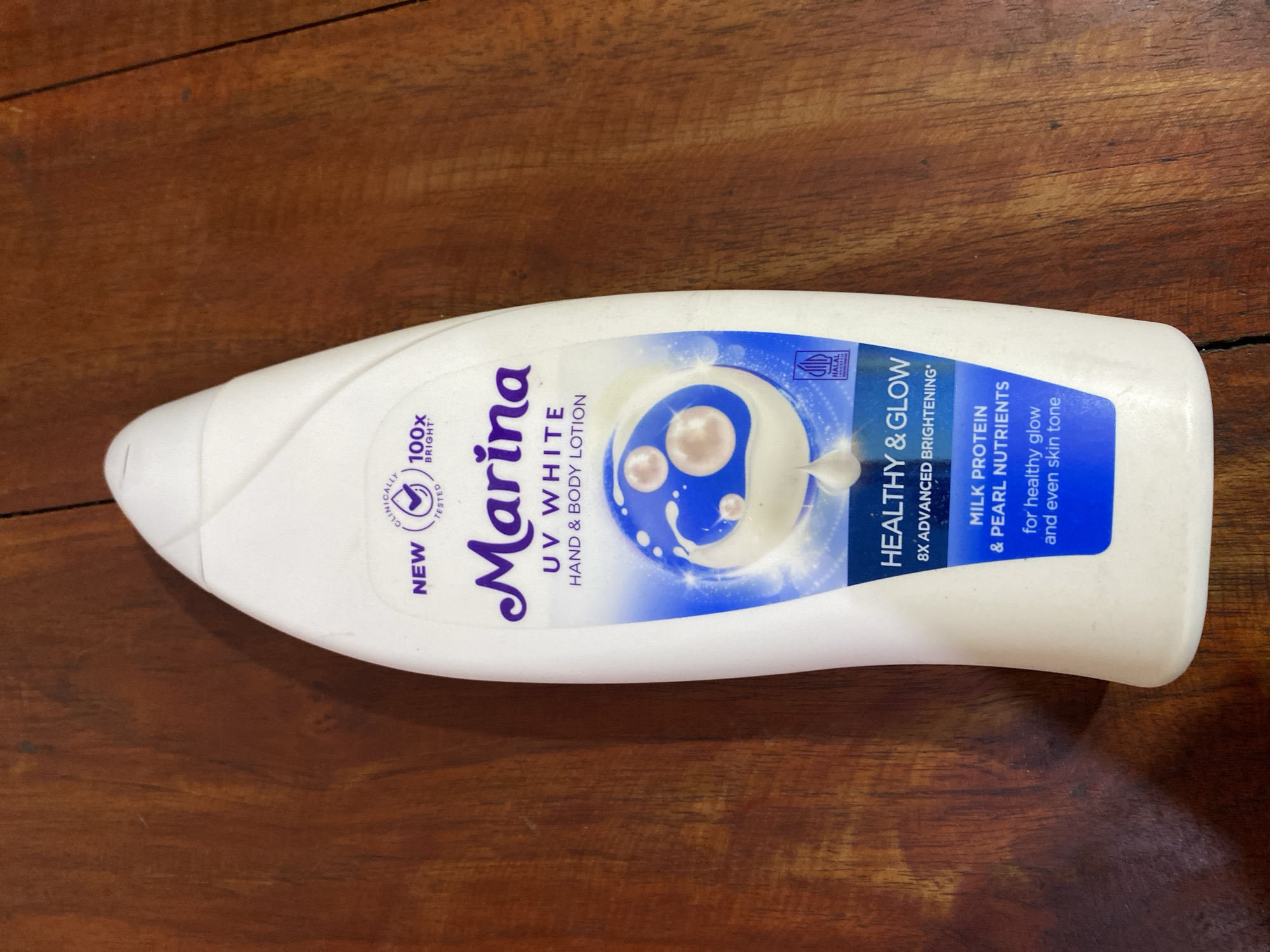 A white plastic bottle of Marina UV White Hand & Body Lotion is displayed on a dark wooden surface. The bottle has a label with text and graphics, including "Marina," "UV WHITE," "HAND & BODY LOTION," "HEALTHY & GLOW," "8X ADVANCED BRIGHTENING*," "MILK PROTEIN & PEARL NUTRIENTS," and "for healthy glow and even skin tone." There are also icons indicating "NEW," "CLINICALLY TESTED," and "100x BRIGHT*." A small "HALAL INDONESIA" logo is visible. The background is a wooden table with visible grain and slight imperfections. There are no people or specific city information visible in the image.