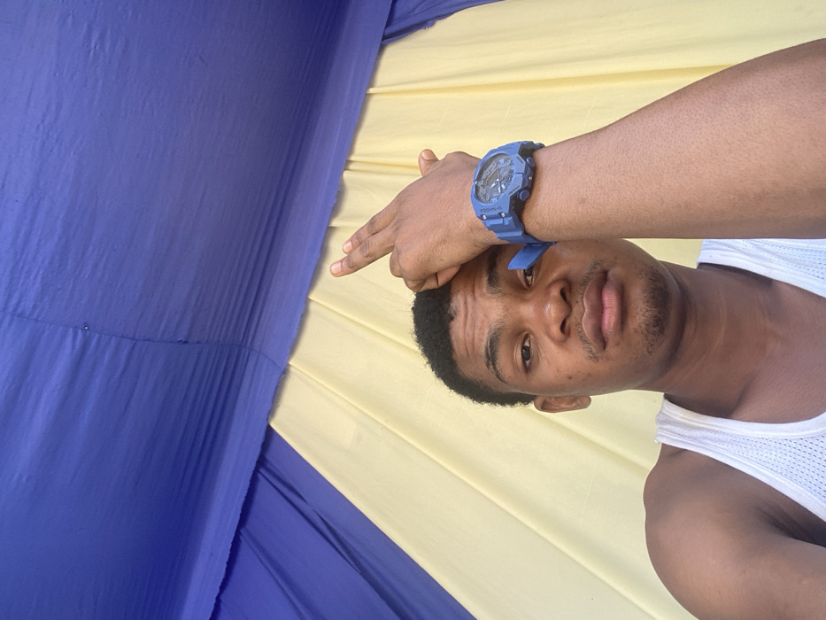A close-up image depicts a male individual, appearing to be a young adult, looking directly at the camera. He has short, dark, curly hair and light stubble on his chin. He is wearing a white mesh tank top. His right arm (viewer's left) is bent, with his right hand resting on top of his head, fingers slightly splayed. A blue digital wristwatch with a robust casing is worn on his right wrist. The background consists of alternating vertical panels of royal blue and light yellow fabric, suggesting an indoor setting with decorative drapes, possibly within a tent or an event space. This scene is located in Ikot Ete Ekpo, Nigeria.