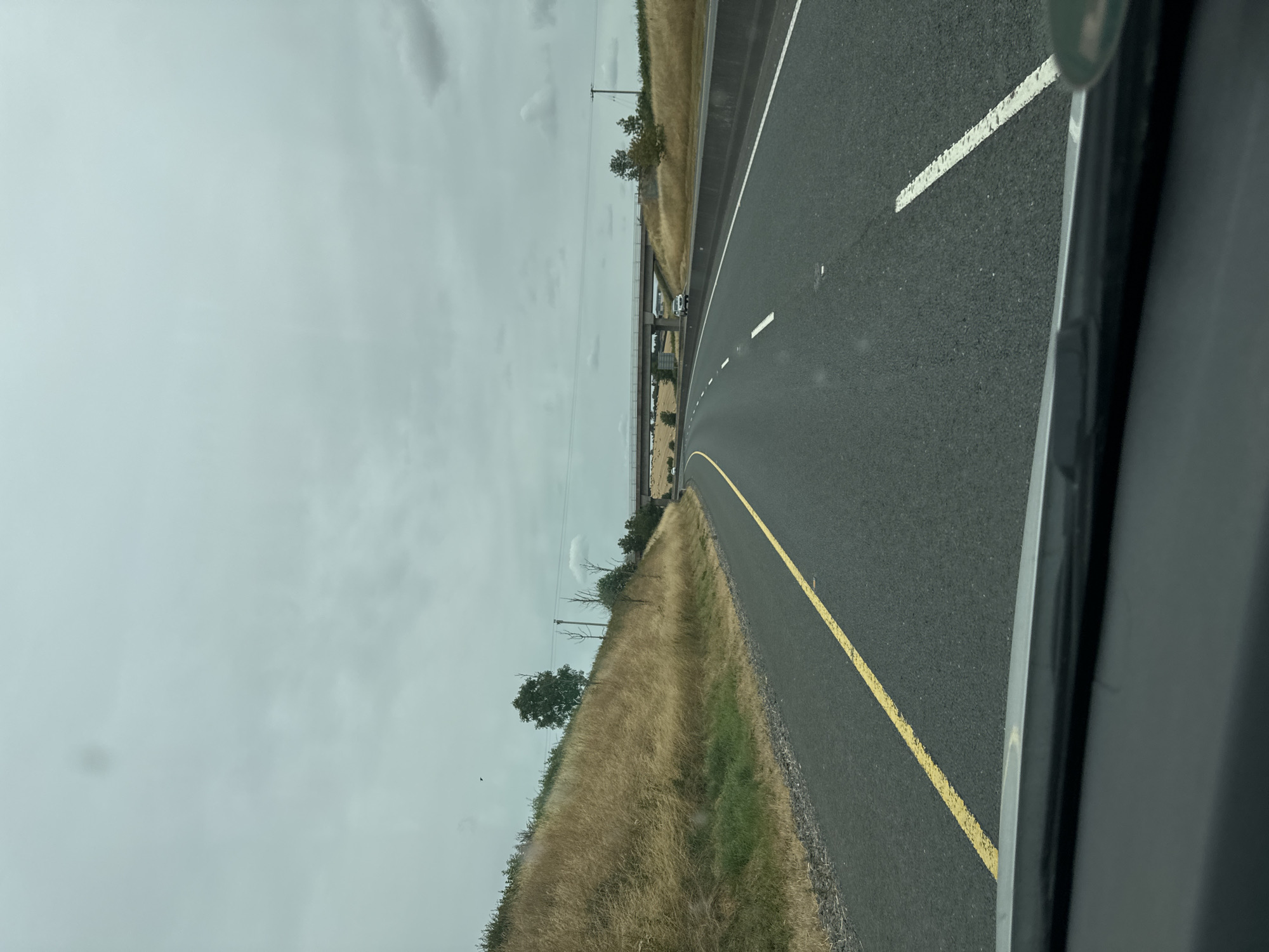 The image depicts a view from inside a vehicle traveling on a highway in the Moyle area of Ireland. The highway is paved with asphalt and marked with a solid yellow line on the left and dashed white lines on the right, indicating multiple lanes of traffic. The right side of the highway is bordered by a concrete barrier. Beyond the barrier, a grassy embankment with dry, golden-colored vegetation rises steeply, dotted with scattered trees and power poles. In the distance, a concrete bridge spans the highway. The sky is overcast with a muted gray hue. A few cars are visible on the highway in the distance. The overall scene suggests a rural or semi-rural highway environment under cloudy weather conditions.