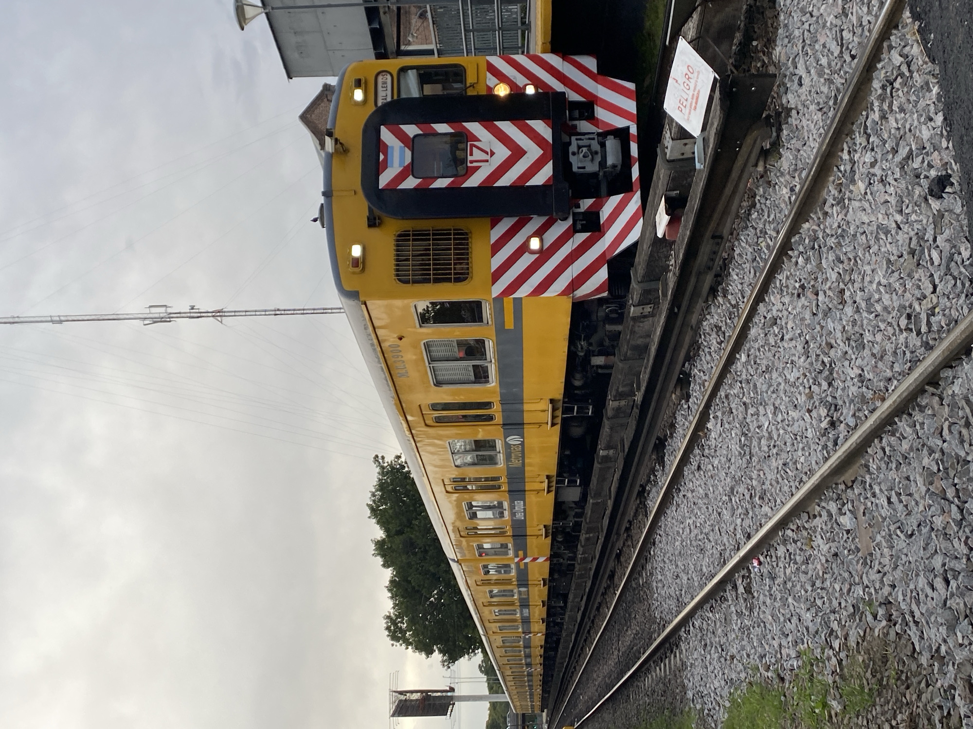 An image captures a bright yellow and black train from a side-front perspective, positioned on railway tracks under an overcast sky. The train appears to be stationary, possibly at a station or siding.
The front section of the train features a striking black panel with bold red and white chevron stripes, which serve as a high-visibility marking. A small sticker of the Argentine flag is visible above the main windshield, and below it, the number "17" is displayed in red on a white background, accompanied by a red chevron pointing to the right. Two rectangular headlights are illuminated. Above the windshield, the text "GRAL. LEMOS" is visible on the yellow body.
Along the side, the train car is primarily yellow with horizontal grey stripes, featuring multiple windows. The words "Metrovías" and "Línea Urquiza" are clearly visible, identifying the operator and the specific railway line. The car number "M.U. 3900" is also legible.
Two parallel railway tracks, covered with grey gravel ballast, extend from the foreground into the distance. Adjacent to the tracks, a white warning sign on a post prominently displays "PELIGRO" (Spanish for "DANGER") in red uppercase letters, with additional smaller text below.
The sky is uniformly overcast and light grey, suggesting a cloudy daytime. In the background, power lines, parts of buildings, and green tree foliage are discernible. The scene depicts a typical railway environment, consistent with a location such as Sáenz Peña, Argentina. No people are visible in the image.