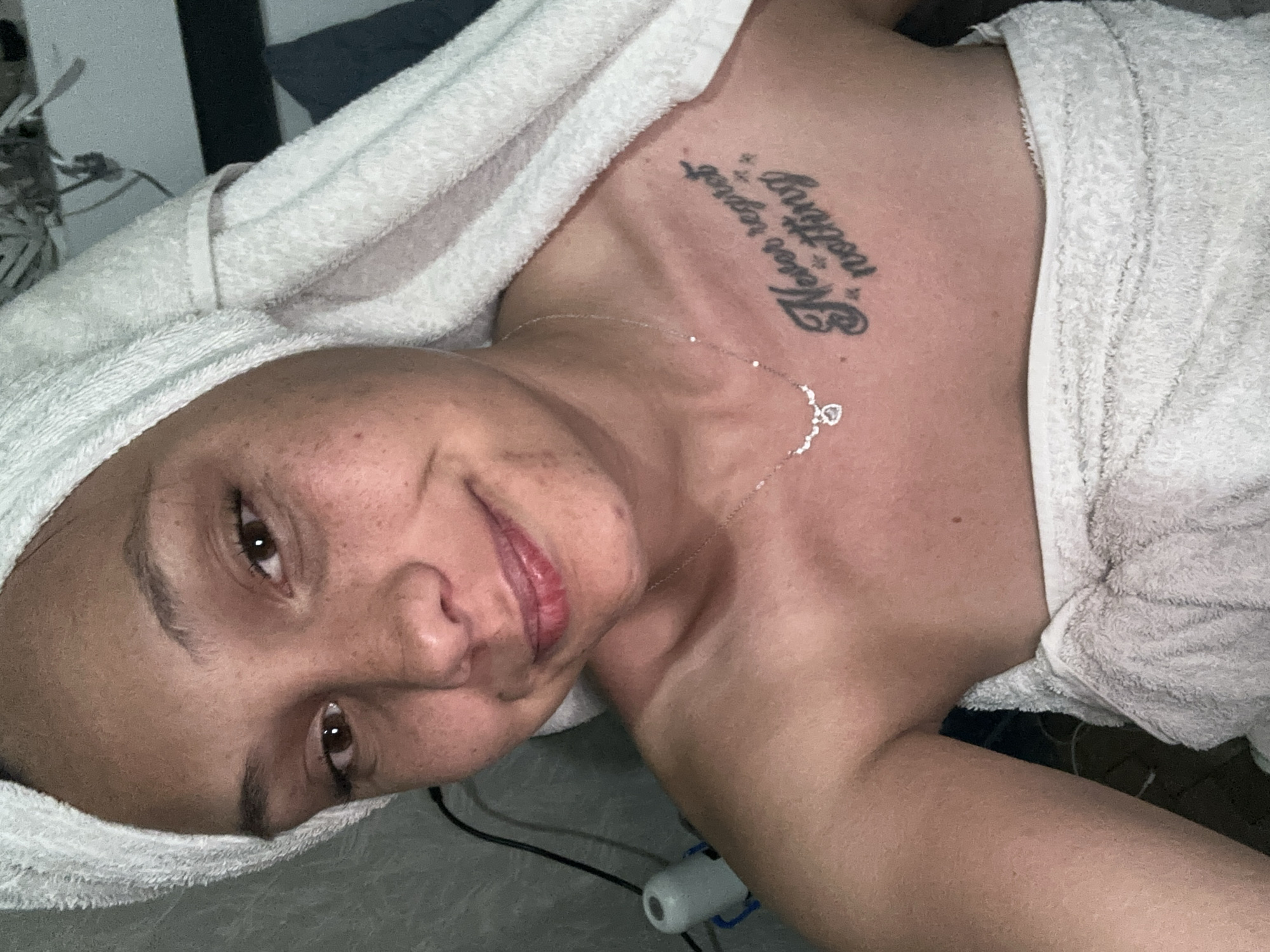 An adult female is captured in a close-up, top-down view, lying on her back and looking directly at the camera with a soft smile. Her head is tilted, and her face is adorned with freckles. She has dark hair, which is completely wrapped in a white towel, suggesting she may have just showered or is preparing for a relaxing activity. Another white towel, possibly part of a bathrobe, covers her upper body.
A delicate silver chain necklace with a small, heart-shaped or similar pendant hangs around her neck, resting on her chest. Prominently displayed on her upper right chest/shoulder area is a tattoo written in a stylized, cursive font that reads: "Never regret nothing."
The setting appears to be indoors, with the person lying on a surface that looks like a bed or a sofa, indicated by the indistinct grey fabric beneath her. In the background, a dark pillow or blanket and some wires connected to a light-colored, cylindrical device are partially visible near her left shoulder. The overall impression is one of a casual, relaxed moment.