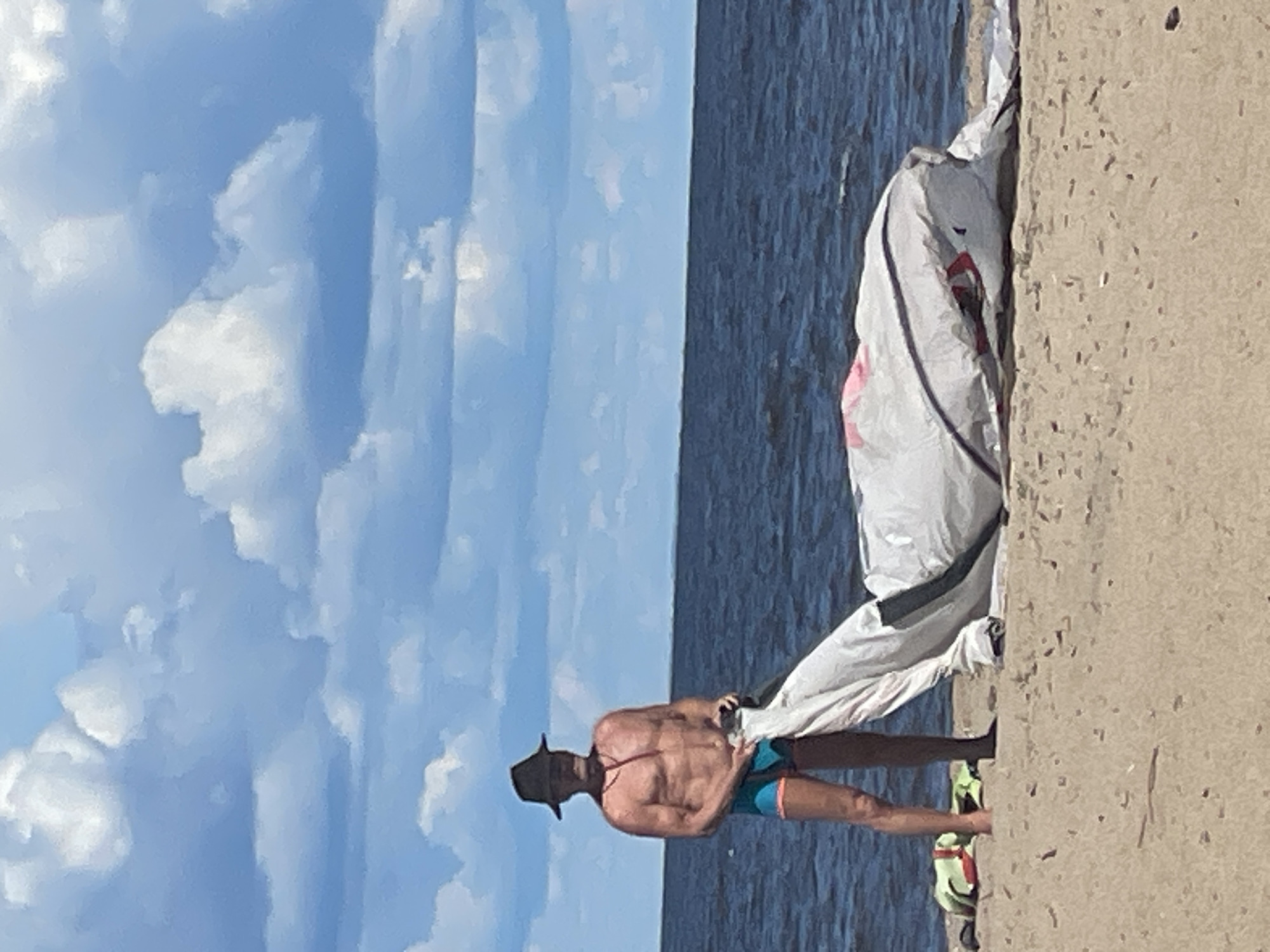 A shirtless man wearing a wide-brimmed hat and swim trunks stands on a sandy beach. He is holding a white and gray object, possibly a kite or sail, which is spread out on the sand in front of him. The man is looking down at the object. The ocean is in the background, with a blue sky and clouds above. The location is the beach in Belozersk, Russia.