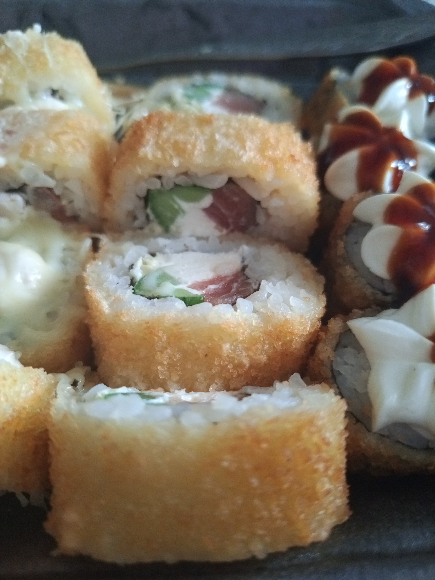 The image shows a close-up of various pieces of sushi, likely served together. The predominant type is a golden-brown, breaded and deep-fried roll, filled with white rice, what appears to be salmon, cream cheese, and avocado. Another type is visible on the right, topped with dollops of white cream sauce and a drizzle of brown sauce. Some sushi pieces have a topping of melted cheese. The scene appears to be indoors. There is no information about Уральское, Russia, in the image.