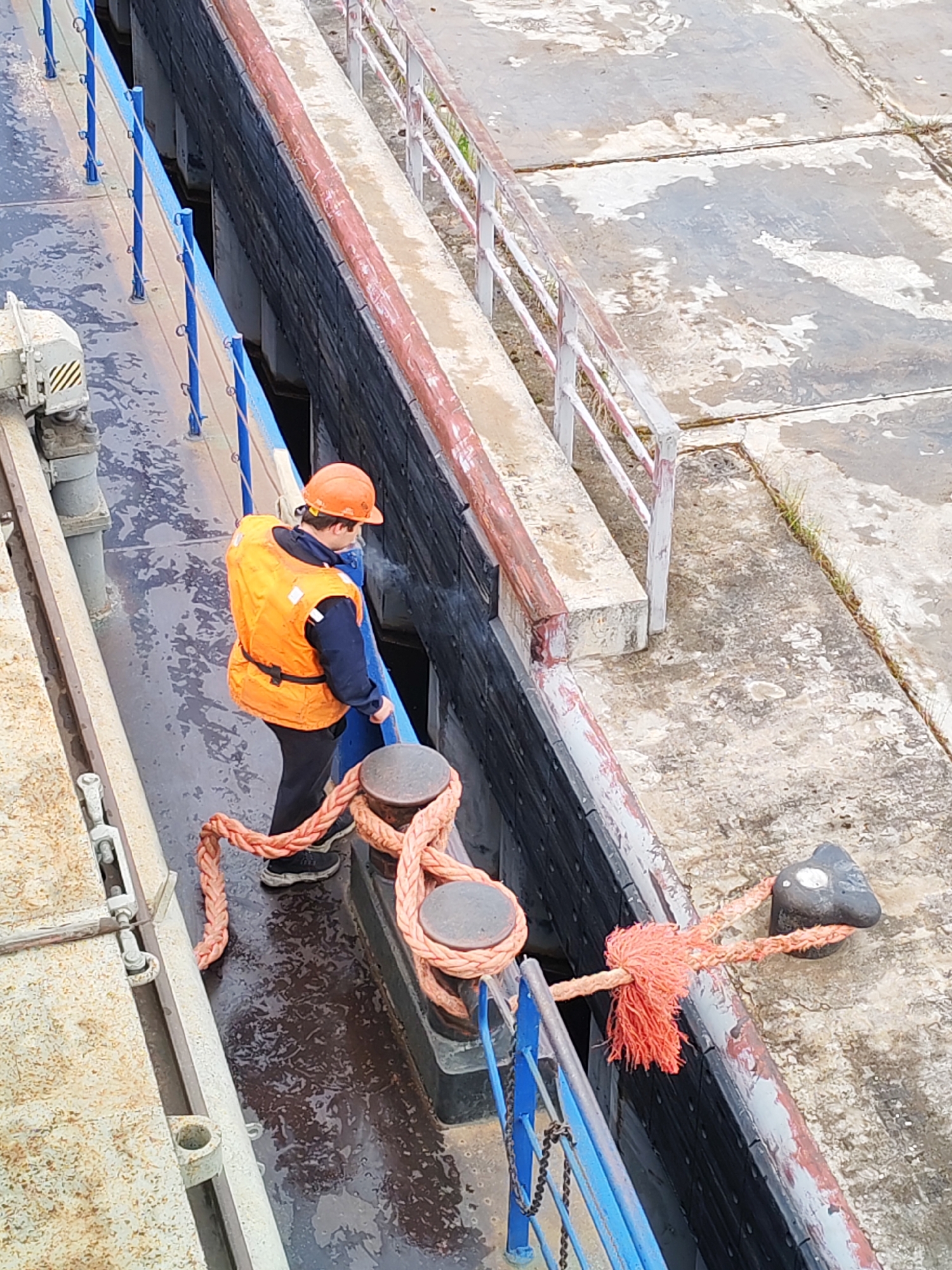The image shows an industrial scene, likely a waterway lock, in Белоусово, Russia. A person wearing an orange hard hat and high-visibility vest is standing on a metal walkway. They appear to be handling a thick, light-red rope that is secured to metal bollards. The walkway has blue railings on one side and a concrete barrier on the other. The ground surface is concrete, showing signs of weathering and moisture. Adjacent to the walkway is a deeper recessed section, possibly part of the lock mechanism. The scene has a functional, utilitarian feel, typical of industrial settings.