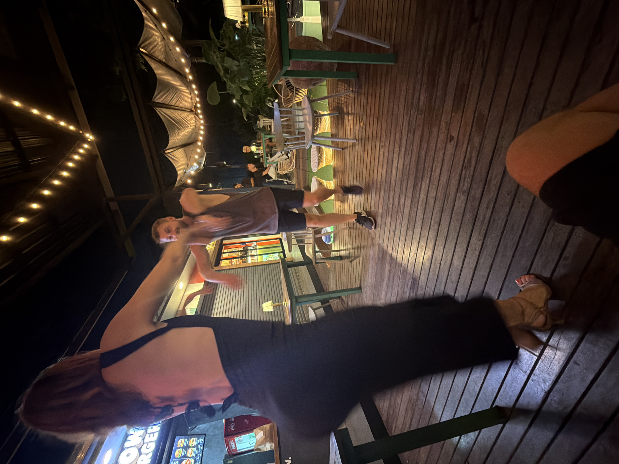 In a dimly lit outdoor setting, likely a restaurant or bar in Ban Rang Rak, Thailand, several people are visible. The scene is a wooden deck with string lights overhead, suggesting an evening event. One person in the foreground is wearing a black dress and has red nail polish. Another person, appearing to be a man, is wearing a grey t-shirt and black shorts, and is captured mid-movement, possibly dancing or exercising. A third person, a woman with red hair, is seen from behind, also in a black outfit. In the background, other individuals and outdoor seating arrangements are discernible, including chairs and tables, amidst lush greenery. A menu board with text, including "OWN BURGER," is partially visible, further indicating a food establishment. The overall atmosphere appears casual and social, with the warm glow of the lights creating an inviting ambiance. The time of day is clearly night, and the weather seems to be warm and pleasant, suitable for an outdoor gathering.