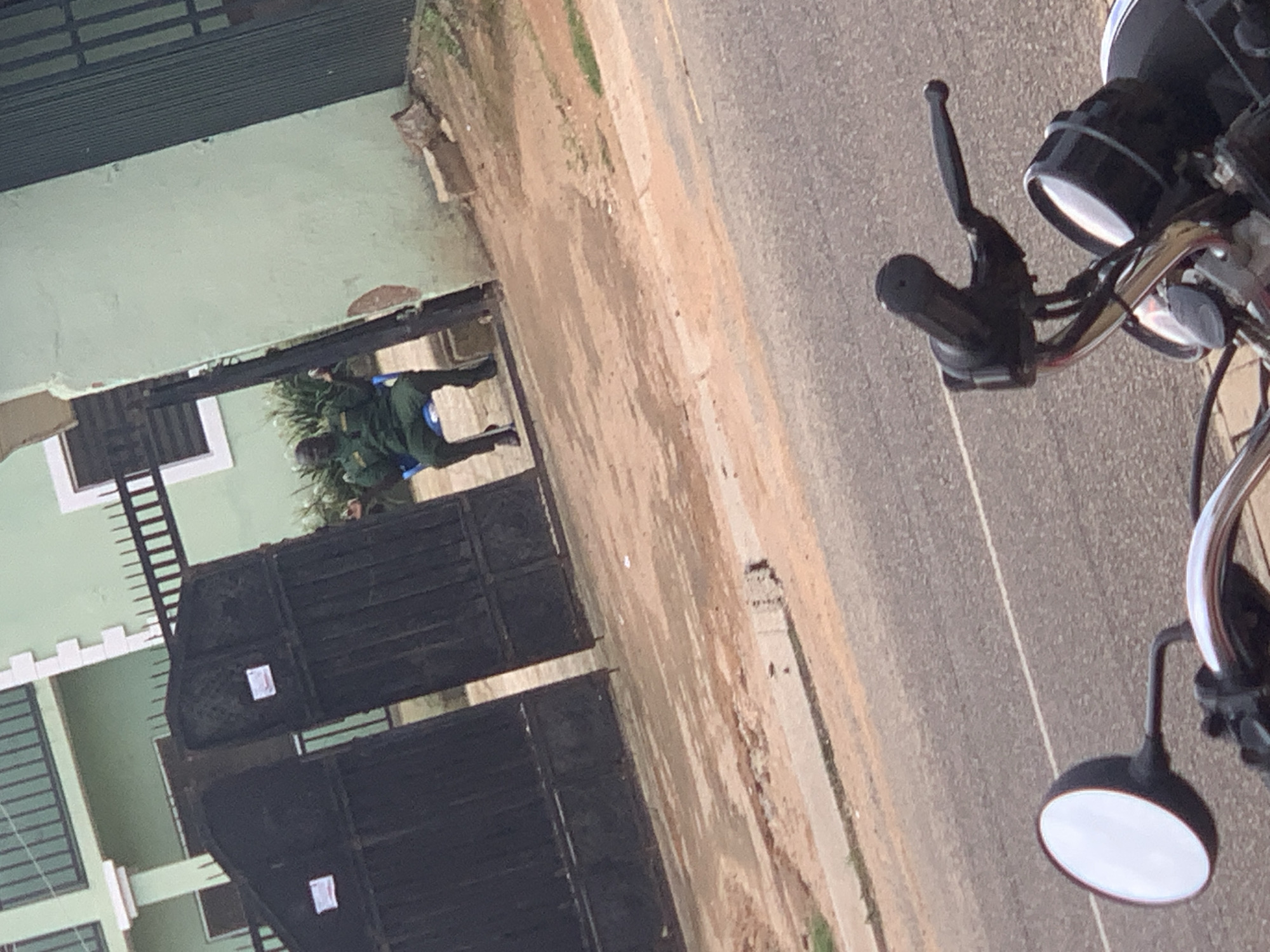 In Jos, Nigeria, two individuals dressed in what appears to be military or security uniforms stand near a large, dark metal gate. The gate is segmented into two main sections and has white square labels on each. Behind the gate, a light green building with white trim and windows is visible. One of the individuals is standing next to a small, blue chair. The scene appears to be outdoors, with a dirt path bordering the paved road. On the right side of the frame, a portion of a motorcycle is visible, including its handlebars, a mirror, and a headlight. The lighting suggests it is daytime, and the weather appears to be clear. There is no visible text other than the labels on the gate.