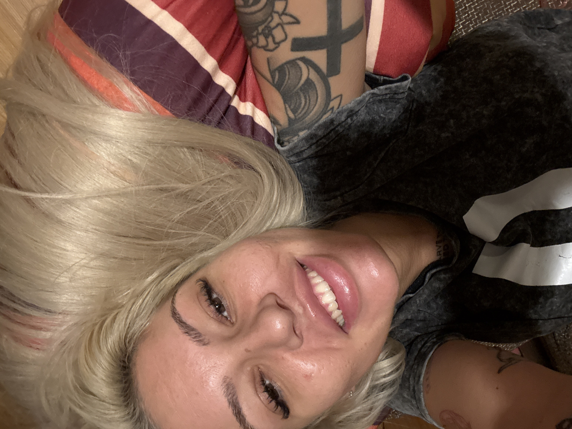A selfie shows a woman with blonde hair lying down. Her head is tilted towards the left. She has fair skin, brown eyes, and is smiling, revealing her teeth. Her right arm is visible, showing tattoos on her upper arm and shoulder. She is wearing a dark grey t-shirt with white stripes. The background includes a striped cushion in shades of red, purple, and white. There is no indication of Plovdiv, Bulgaria or any other city.