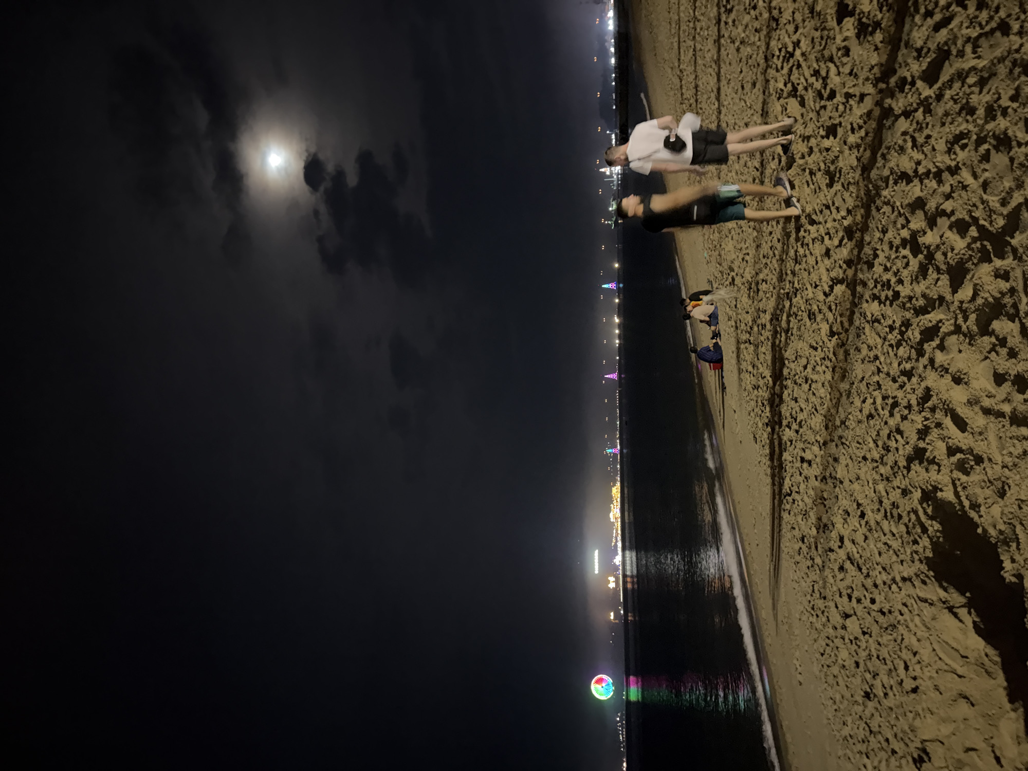Two people are on a sandy beach at night. One is wearing a white shirt and black shorts; the other is wearing a black shirt and blue-green shorts. Behind them are other people on the beach. The city of Nha Trang Ward, Vietnam, can be seen in the distance with its lights reflecting off the water. The moon is visible through the clouds in the sky.