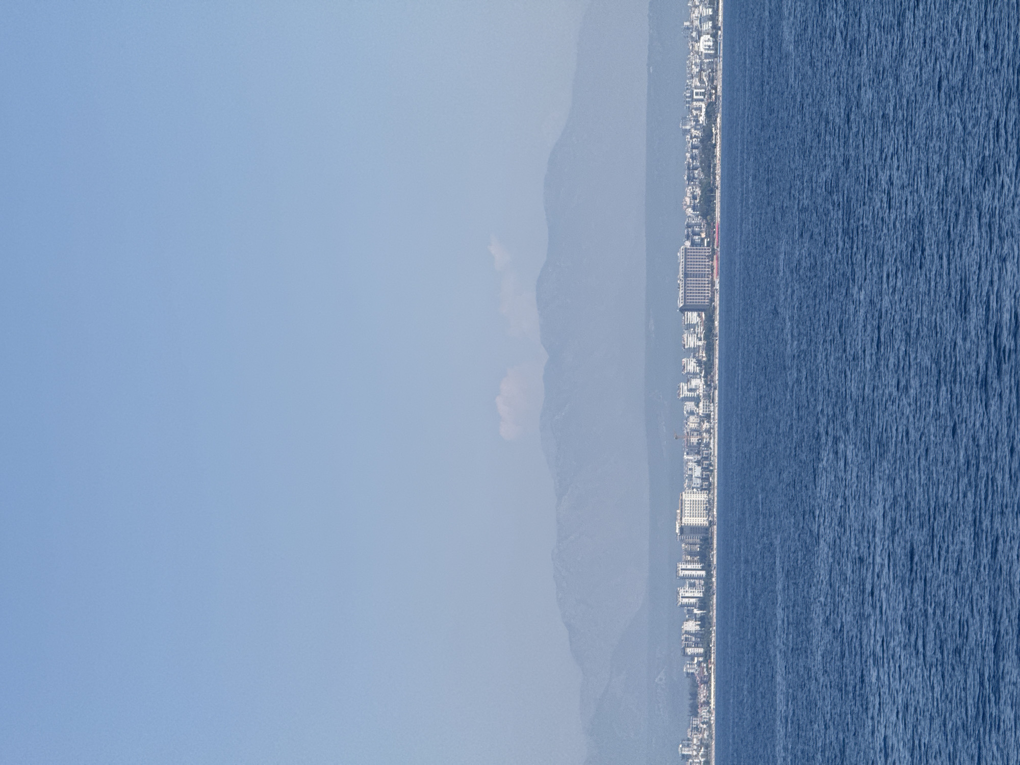 An elevated, wide shot captures the coastline of Konyaaltı, Turkey, on a clear day under a pale blue sky. The foreground is dominated by the deep blue expanse of the sea, its surface rippled by gentle waves. In the midground, a strip of cityscape lines the shore, characterized by a dense collection of mid-rise buildings and a few taller, modern structures, including what appears to be a large hotel with a distinctive facade. The buildings are tightly packed, forming a continuous urban line. Beyond the city, a range of hazy, blue-toned mountains rises in the background, their peaks softened by atmospheric perspective. A few wisps of clouds are visible high in the sky. The scene depicts a coastal city situated against a mountainous backdrop, with the sea stretching out before it.