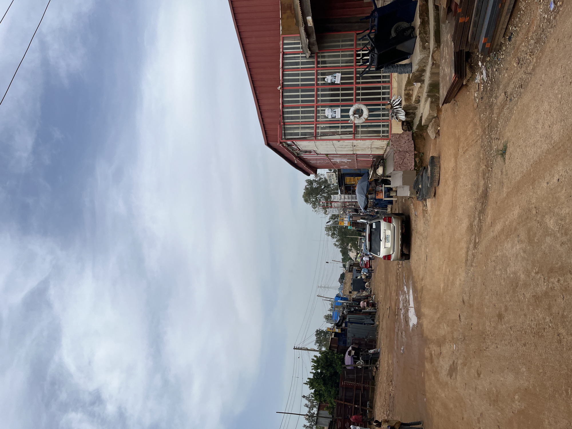 The image depicts an outdoor scene in Gura Top, Nigeria, likely in a marketplace or commercial area. The setting appears to be a street with a dirt and gravel ground, bordered by buildings and various stalls.
In the foreground, a tan-colored SUV with a license plate that reads "KJA 670H" is parked on the right side of the road. The ground in front of it has puddles, suggesting recent rain. A few people are visible near the car and further down the street.
To the left of the road, a red-roofed building with large windows is prominent. Some posters are attached to the windows, and a white life preserver ring is hanging on the facade. A dark-colored wheelbarrow is also visible near the building. Further down the street, more stalls and structures are lined up, with some displaying blue tarps or corrugated metal roofs. Trees are scattered throughout the scene, indicating a semi-rural or suburban environment.
The sky is overcast with scattered clouds, suggesting a possibly cloudy or humid day. The overall impression is one of a bustling, somewhat unkempt commercial area. The time of day appears to be daytime, given the ambient light. There is no obvious indication of a specific event, but rather a snapshot of daily life in this location.