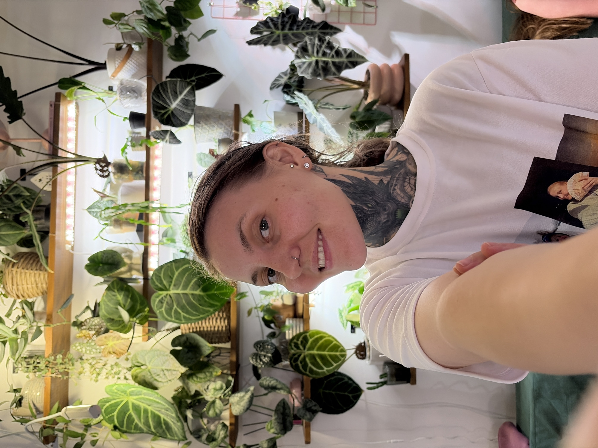 The image shows a young woman with a large tattoo on her neck and arm, taking a selfie. She is wearing a white t-shirt with a printed image. She is standing in front of a wall filled with numerous potted plants on shelves. The shelves are illuminated by LED grow lights. The environment appears to be indoors, possibly a home, with a plush green seating area visible in the foreground. The location is stated to be Malyi Bukor, Russia.