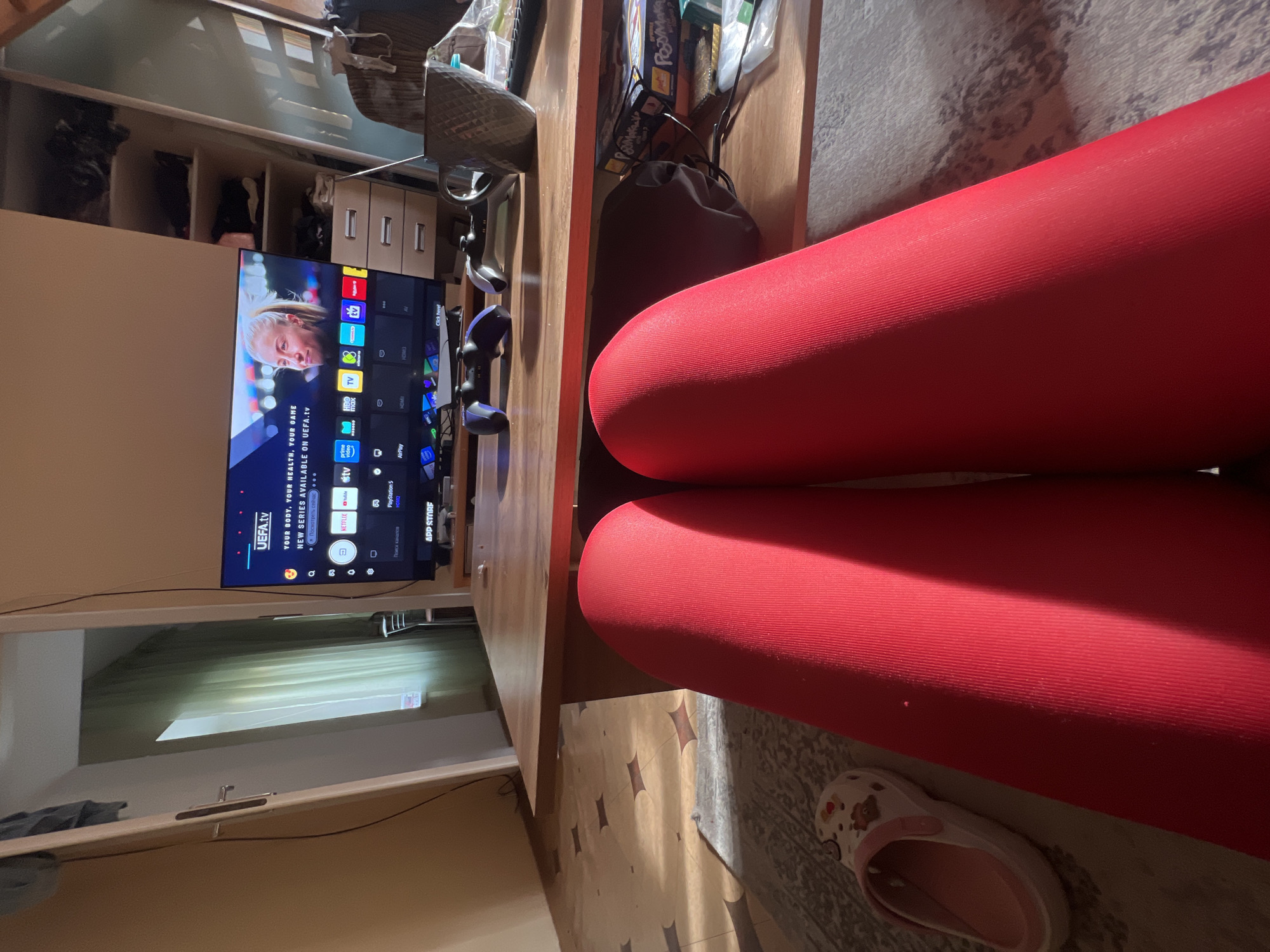 The image captures an indoor scene from a first-person perspective, looking towards a television screen. The lower right foreground is dominated by two prominent red cylindrical shapes, likely rolled-up yoga mats or large cushions, under which a light-colored slipper or Croc rests on a patterned rug.
A smart television is visible on a wooden desk, displaying a menu interface. The UEFA.tv app is highlighted, with text reading "YOUR BODY, YOUR HEALTH, YOUR GAME" and "NEW SERIES AVAILABLE ON UEFA.tv." Other visible app icons and text include HBO TV, Netflix, Apple TV, Prime Video, YouTube, PlayStation 5, AirPlay, and App Store. Text like "Посмотреть сейчас" (Watch now) and "Поиск к" (Search to) indicates the interface is partially in Russian.
On the wooden desk, two gaming controllers, consistent with PlayStation 5, are placed alongside a dark, textured mug. Behind the desk, a shelving unit or cabinet holds various items, including a box with the word "PosMaka" visible. To the left of the television, a window with light-colored blinds allows some light into the room. The overall atmosphere suggests a casual, leisure activity, likely watching TV or gaming, in an indoor setting.