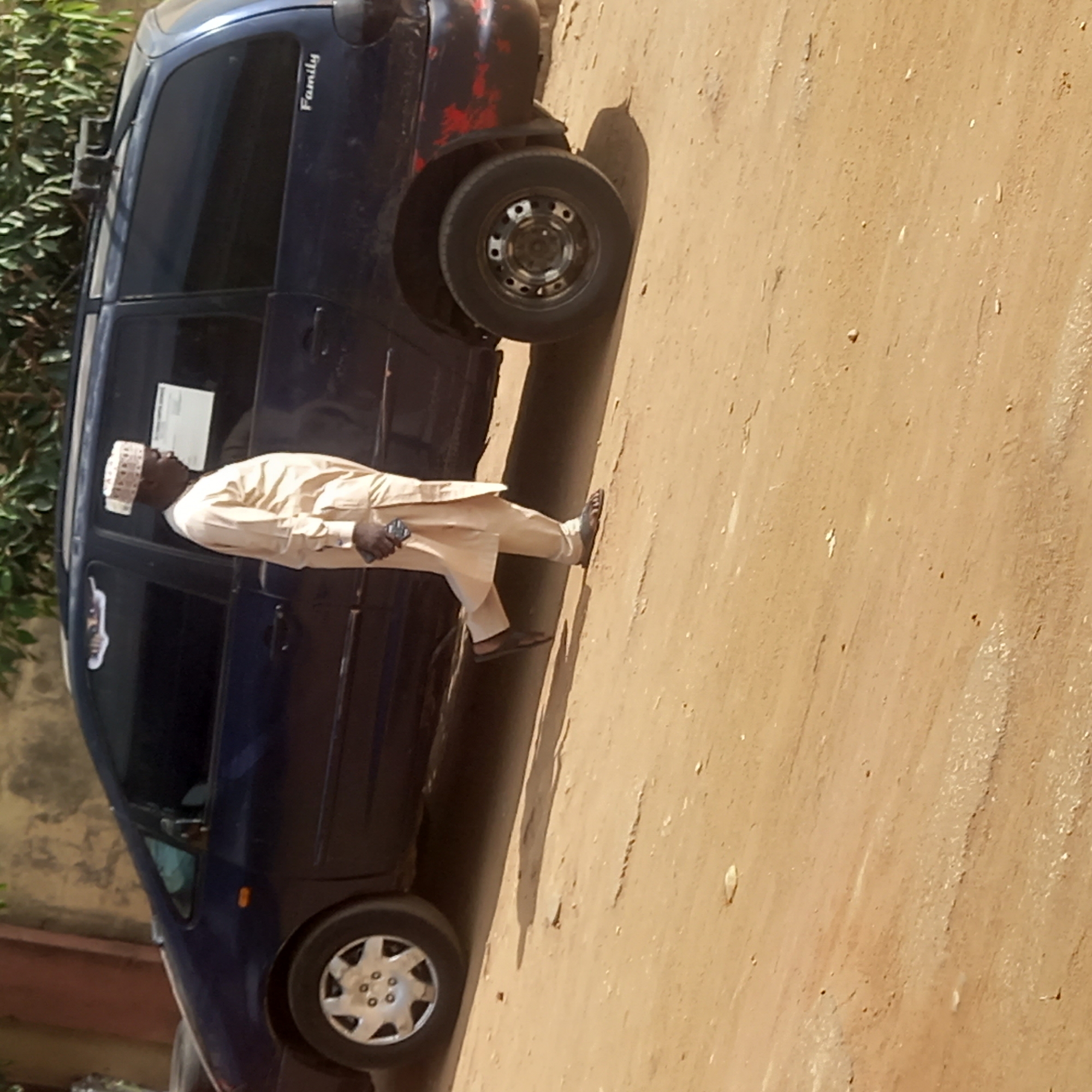 A man in traditional clothing stands in front of a blue minivan with "Family" written on the side, parked on a dirt road in Dadi Riba, Nigeria. The minivan has noticeable wear and tear. The man is dark-skinned, wearing a patterned white cap, light-colored robes, and sandals. He is holding an object in his left hand. The background includes sparse greenery and the sandy-colored road.