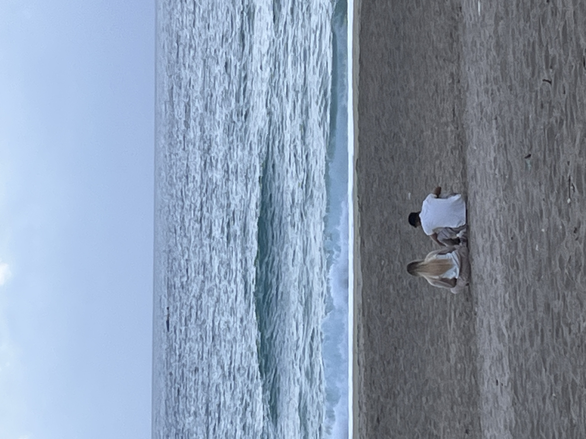 A slightly blurred, vertically-oriented, outdoor photograph shows a couple kneeling on the beach next to the ocean on a cloudy day. The ocean spans the left portion of the image. White-capped waves are visible. To the right of the ocean is the sandy beach with light and dark shadows. A man and woman kneel together on the beach, facing right. The man wears a white t-shirt, tan shorts, and has dark hair. The woman has long blonde hair and wears a white shirt and tan shorts. The location appears to be a beach in Alanya, Turkey.