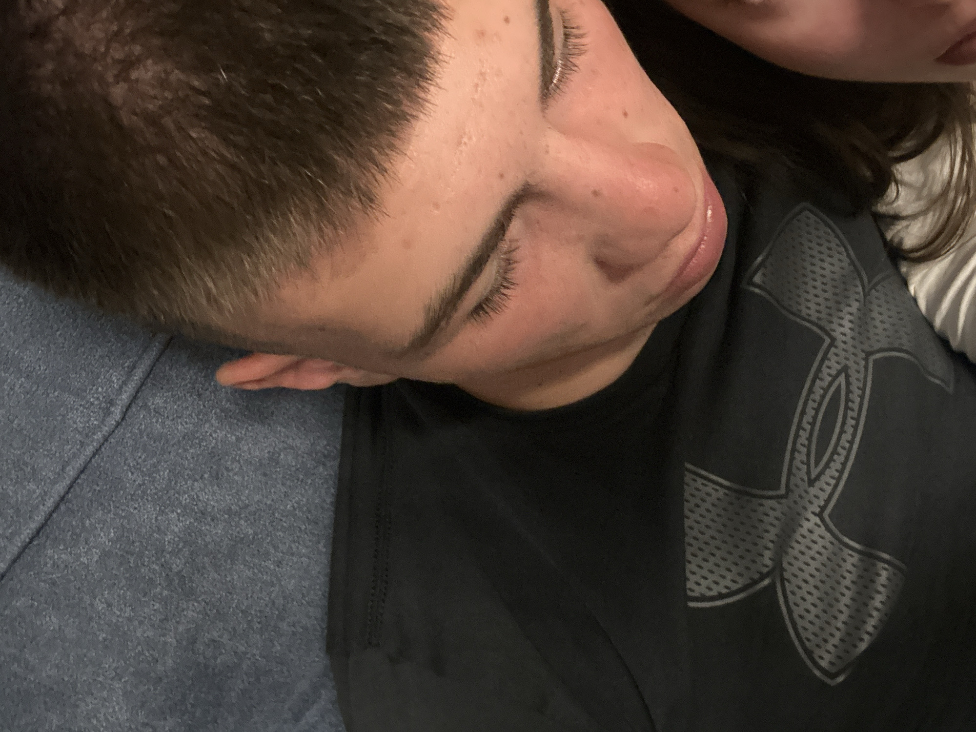 A young person with short, dark brown hair is lying down on a blue textured surface, possibly a sofa or blanket. They are wearing a black t-shirt with a prominent grey Under Armour logo on the chest. Their head is tilted downwards, with their eyes closed or looking away from the camera. Their facial features are visible, including eyelashes and skin with some blemishes. Another person's face is partially visible on the right side of the frame, with their cheek and mouth showing. The lighting suggests an indoor setting, likely at night or in a dimly lit room. There is no indication of the specific location in Sofia, Bulgaria.