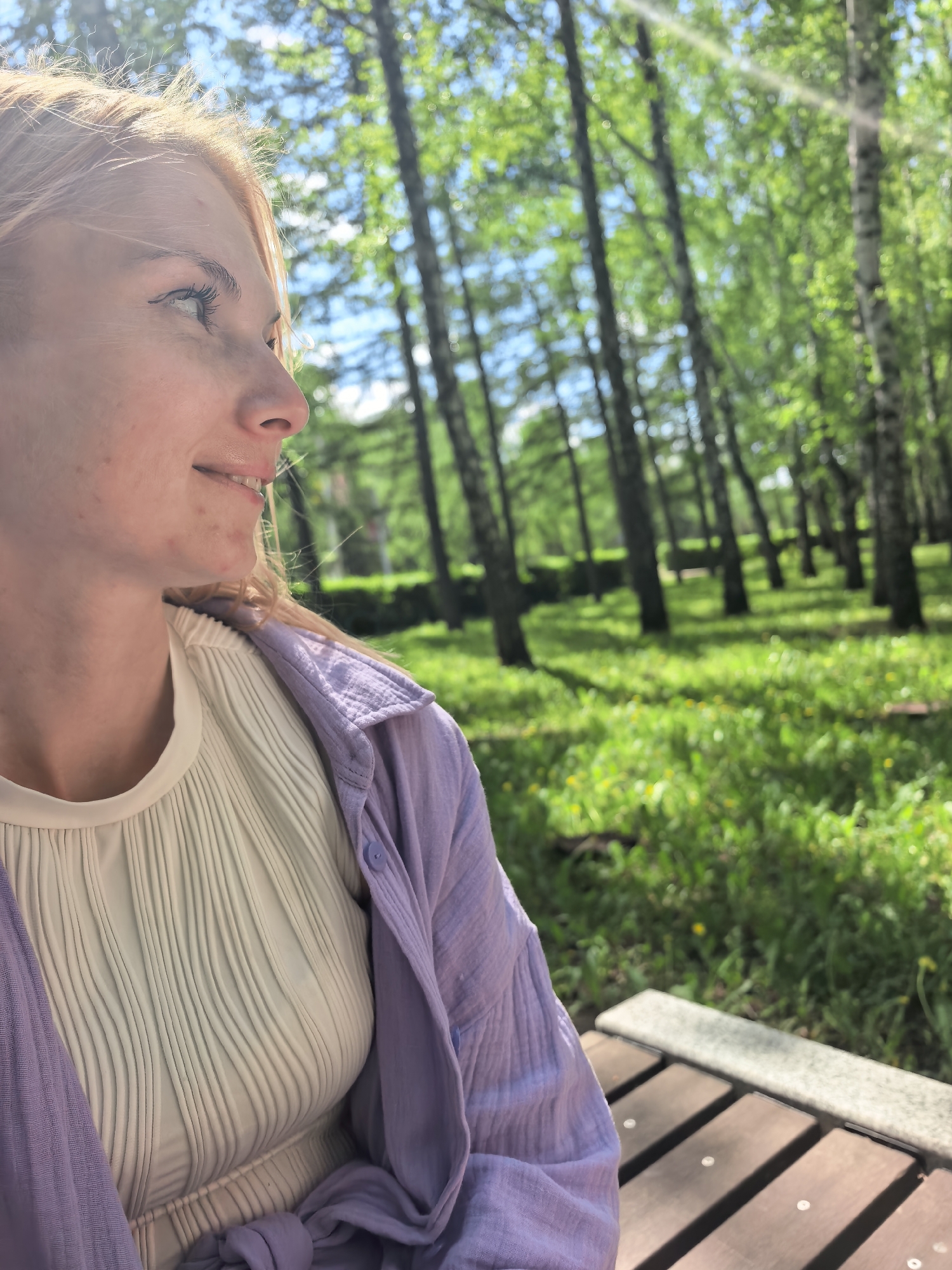 A woman with fair skin and light reddish-blonde hair is sitting on a park bench. She is wearing a white shirt with vertical ribbed detailing and a light purple, button-down shirt over it. Her head is turned to the side, and she is looking up and to the right. Her expression is pleasant. The background consists of a park with many slender trees and green grass. The environment appears to be illuminated by bright sunlight. The bench is made of wooden slats with a grey concrete border. The location is likely Sterlitamak, Russia, based on the park's characteristic trees and general atmosphere.