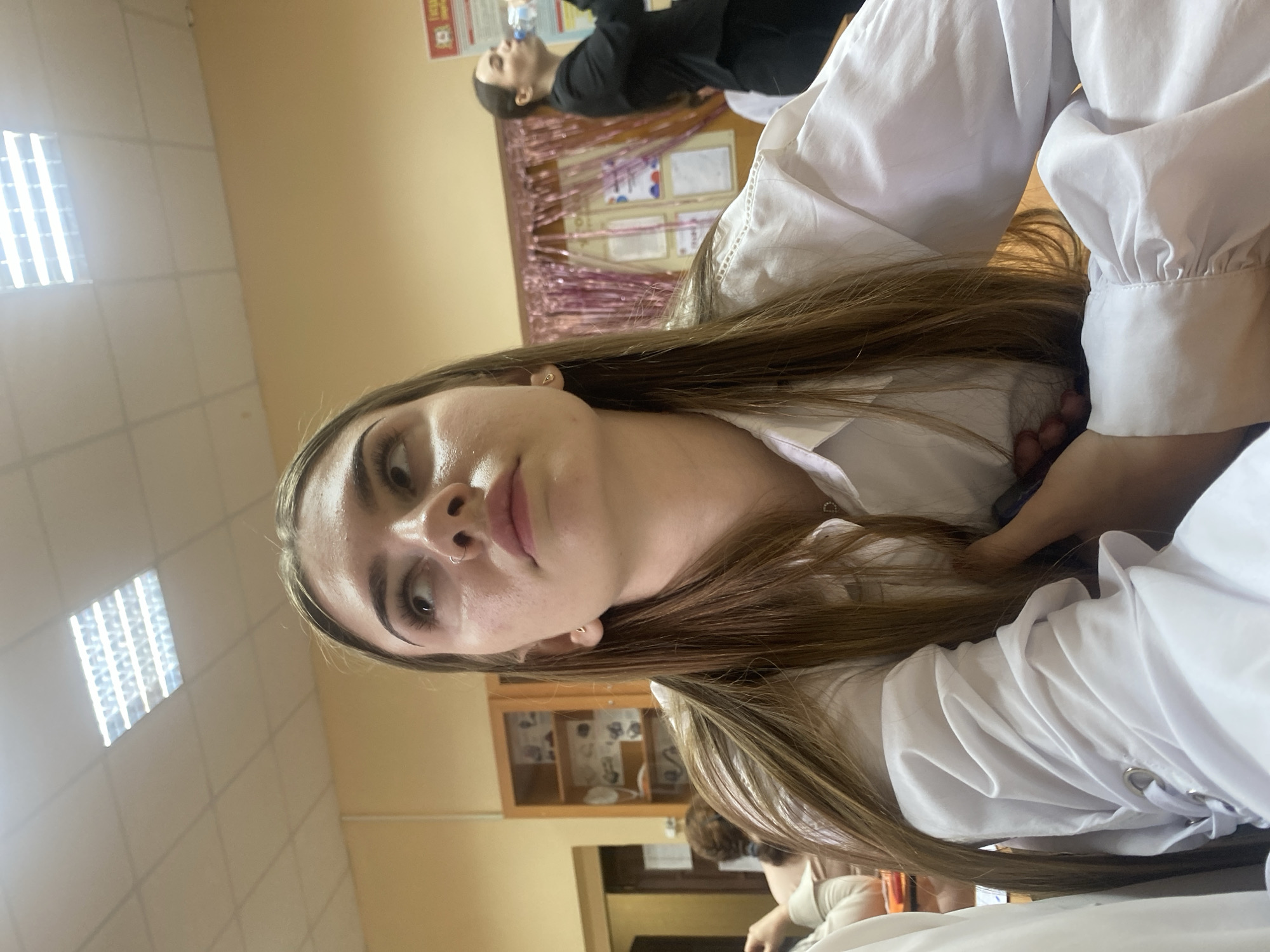 In a classroom setting in Dyakovo, Russia, a young woman with long, brown hair, wearing a white collared shirt, is positioned in the foreground. She is looking upwards and to the right, with her head tilted. Her facial features include styled eyebrows and a nose ring. To her right, another young woman in a black jacket and pants is visible, drinking from a water bottle. In the background, shelves with various items are seen, along with a wall decorated with pink tinsel and posters. The room is illuminated by fluorescent ceiling lights.