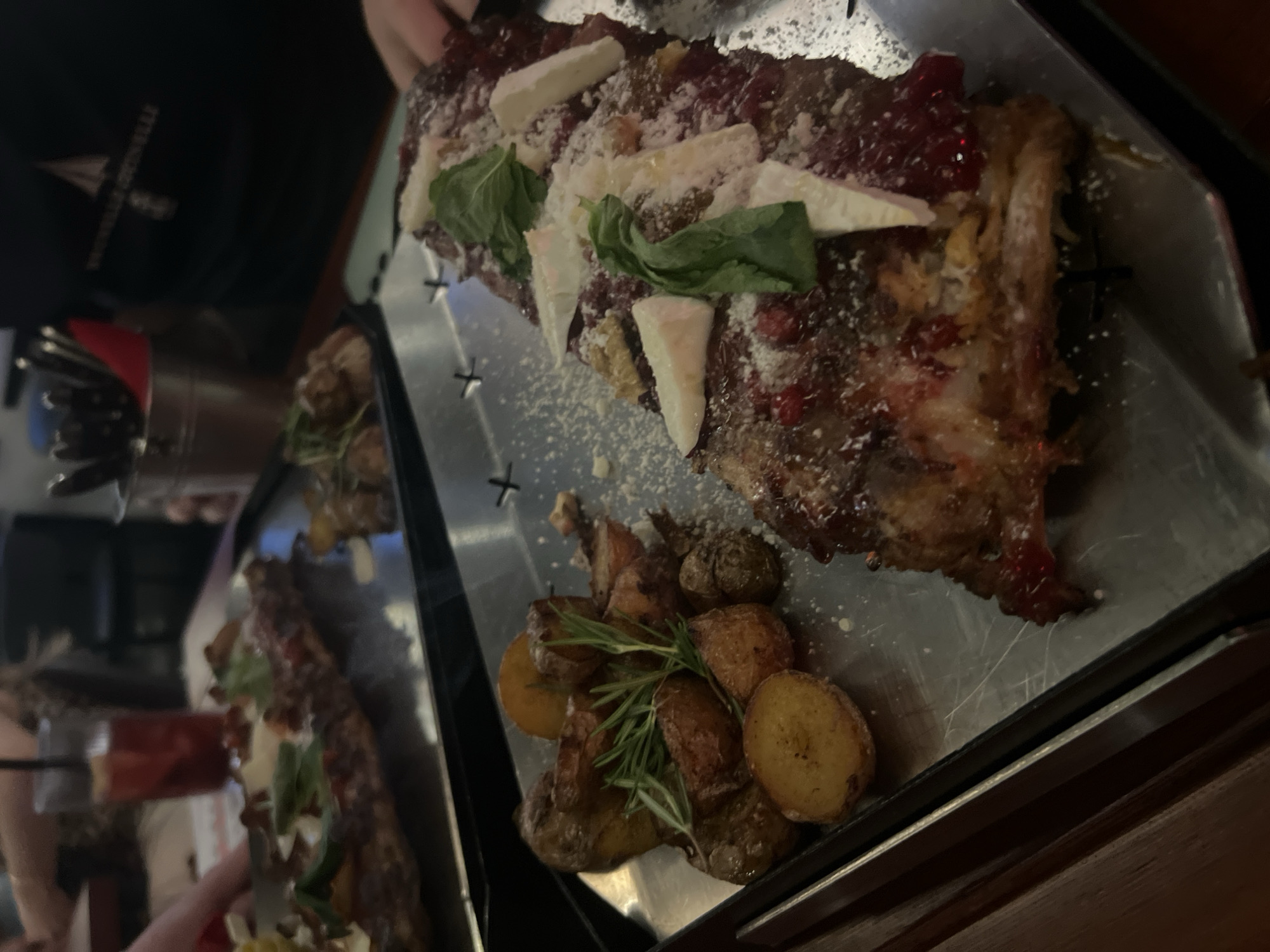 The image displays a close-up view of a large platter of food, likely served in a restaurant during the evening. The main dish features a substantial portion of what appears to be meat, possibly ribs, generously coated in a rich, dark red sauce that contains visible red berries, suggesting a fruit-based glaze like cranberry or lingonberry. The dish is topped with a sprinkle of grated white cheese, several wedges of a soft white cheese (such as brie or goat cheese), and fresh green basil leaves.
Accompanying the main dish is a side of roasted potatoes, which include both halved and whole baby potatoes, garnished with fresh rosemary sprigs. The food is presented on a rectangular metal serving platter that has distinctive cross-shaped perforations.
In the background, a second, similar platter with food is partially visible, hinting at multiple dishes or shared dining. A person's arm and hands are visible on the left, near the background platter. In the upper left corner, part of a dark-colored garment, possibly a shirt or apron, is seen, bearing a white logo with a stylized sail or ship icon and partially legible text that appears to read "TERRA...VELLE", possibly "TERRAVELLE".
The lighting is dim and warm, typical of an indoor dining establishment. The scene captures a moment of dining, emphasizing the artful presentation of the meal. The event is a social gathering or meal at a restaurant, potentially in Krasnoyarsk, Russia.