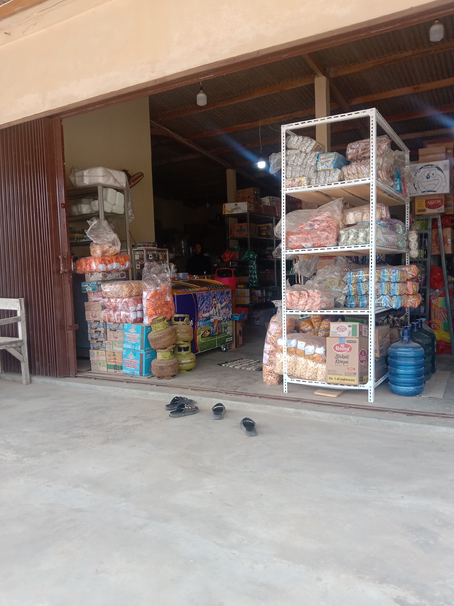 This is a medium-shot, eye-level, outdoor photograph of a small convenience store, likely a warung, in Utama Jaya Mataram, Indonesia. The store is open to the street, with a dark brown metal roll-up door partially open on the left. In front of the store, on the concrete ground, are stacks of merchandise. To the left, there's a small white wooden chair.
The merchandise is diverse and includes numerous packaged food items, such as boxes of biscuits, bags of snacks, and bottles of water. Specifically, there are several large blue water jugs on the right side of the frame. Stacked high on the left are boxes of what appear to be snacks and beverages. In the center, near the entrance, are stacked gas cylinders, a common sight at such stores. A refrigerator with an image of a football player on it is visible behind some of the merchandise.
The interior of the store, visible through the open entrance, shows shelves stocked with more packaged goods. A person in dark clothing is partially visible behind a counter in the back. The lighting is natural, with some artificial lights visible inside the store. The overall impression is of a busy, functional neighborhood shop. A pair of black sandals and a single black sandal are placed on the concrete in the foreground.