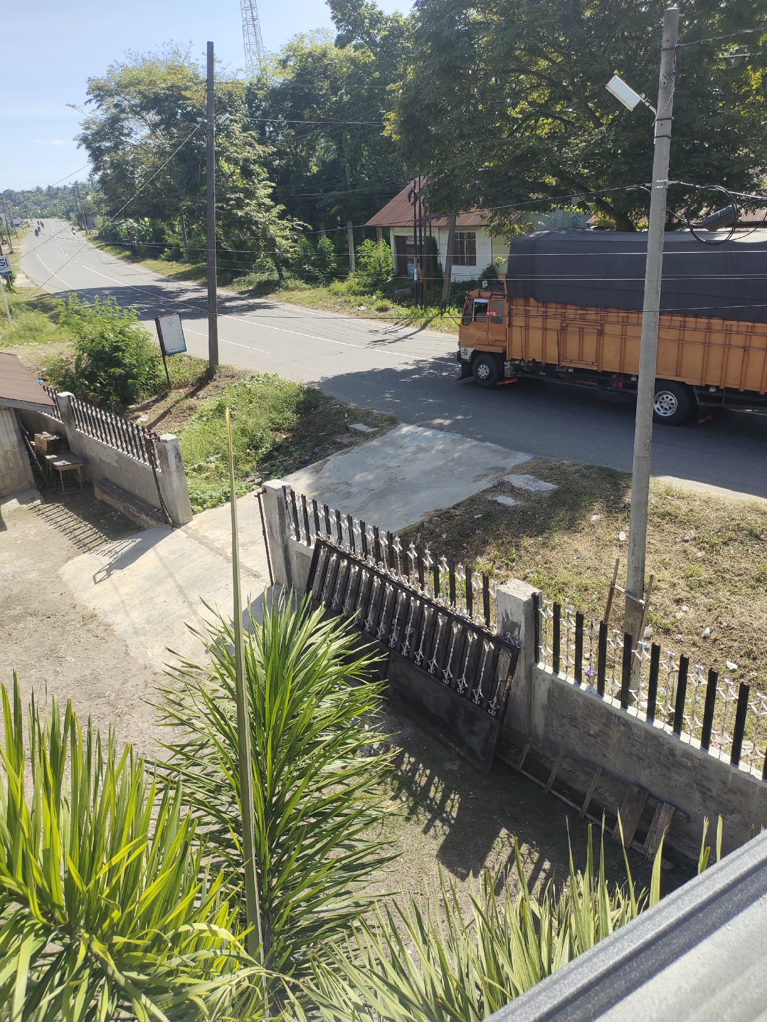A sunny, daytime photograph shows a roadside scene in Meunasahaluedrienlhoksukon Barat, Indonesia. A two-lane asphalt road winds through a residential area with houses and trees. An orange truck with a dark covering is traveling along the road. The view is from an elevated position, looking down at a fence with foliage in the foreground. A utility pole stands near the roadside. The sky is clear.