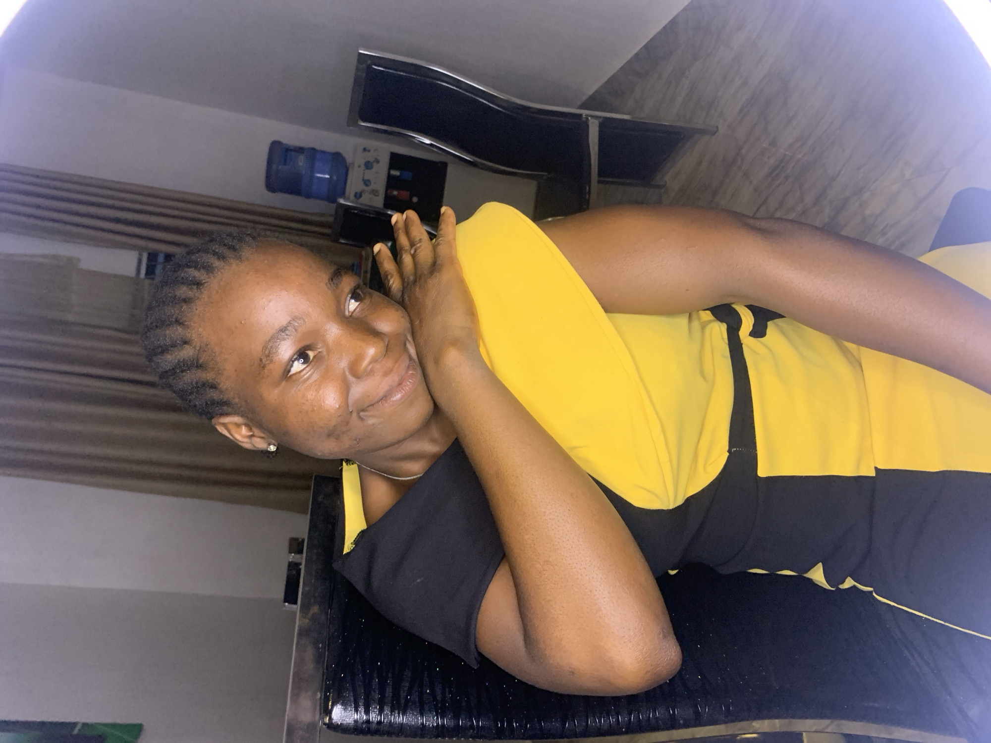 A woman with dark skin and short, braided hair is lying on a black, shiny surface. She is wearing a yellow and black dress. Her head rests on her hand. Behind her is a water dispenser and a black shelf. The location is in Jos, Nigeria.