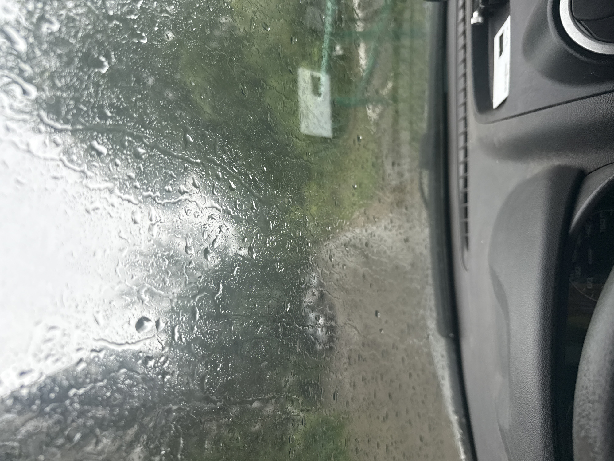 The image shows a view from inside a vehicle looking out through a rain-streaked windshield. The exterior scene is blurred due to water droplets on the glass, but appears to be a rural or semi-rural road lined with trees. A white sign with dark lettering is visible on the right side of the frame, partially obscured by rain. The dashboard of the vehicle is visible in the foreground, with the steering wheel on the left. There are no people or animals visible in the frame. The image was taken on a cloudy or overcast day, consistent with rainy weather. Without further information, it is not possible to determine the specific location within the city of Samara, Russia.