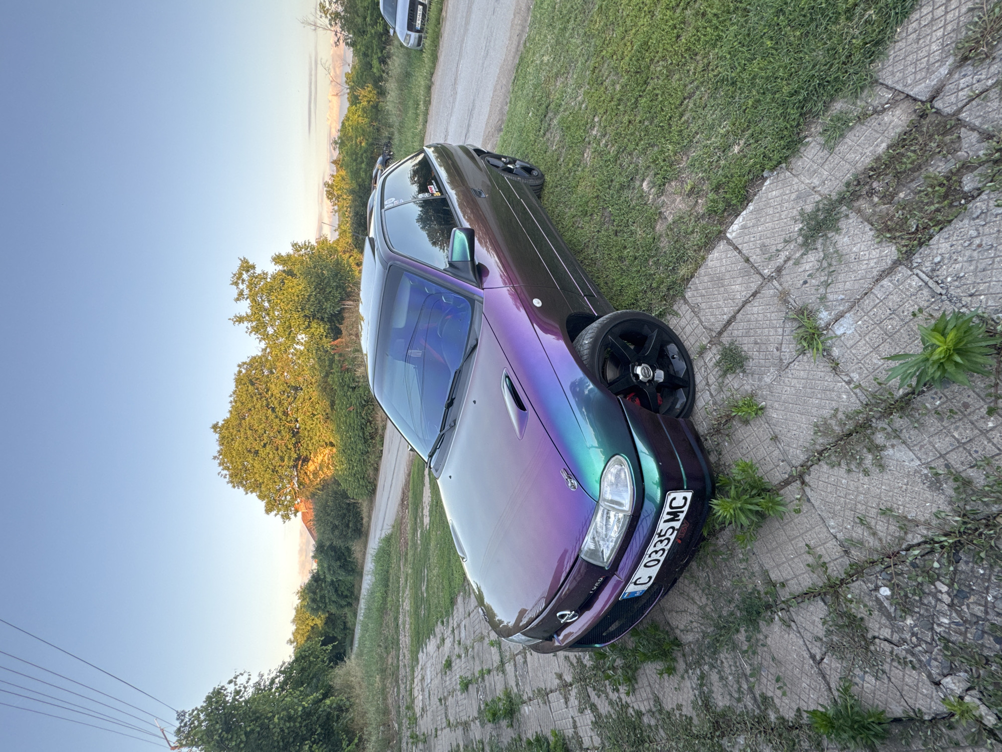 The image shows a custom purple and green iridescent car parked on a cracked concrete path with grass growing through it, bordering a dirt road. The car is a hatchback with black rims and a prominent license plate that reads "C 0335 MC". In the background, there are trees, a building with a red roof, and other vehicles on the road. The sky is clear blue with a hint of sunset colors on the horizon. The overall scene suggests a rural or semi-rural setting, likely in or near Lepitsa, Bulgaria, given the license plate format and the presence of a structure that could be a house.