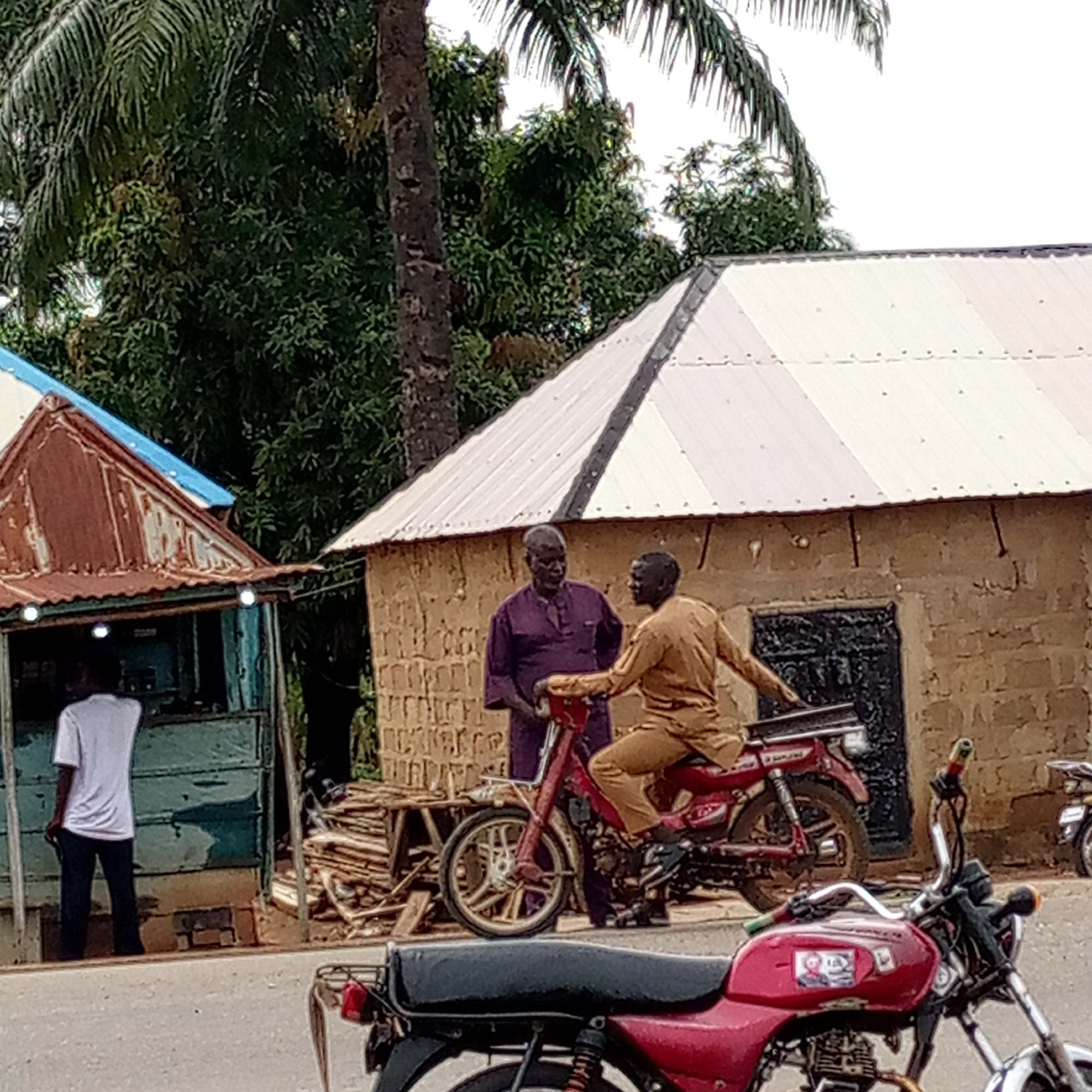 The image depicts a street scene in Mararaba, Nigeria. In the foreground, a red motorcycle is parked on the side of the road. Behind it, a man sits on another red motorcycle while engaging in conversation with a second man who is standing. The standing man is wearing a dark purple outfit. To the left, a person in a white t-shirt and dark pants stands in front of a small, dilapidated blue building. Behind the men and the motorcycles is a tan building with a white roof. A pile of lumber sits between the two buildings. Tall trees are visible in the background.