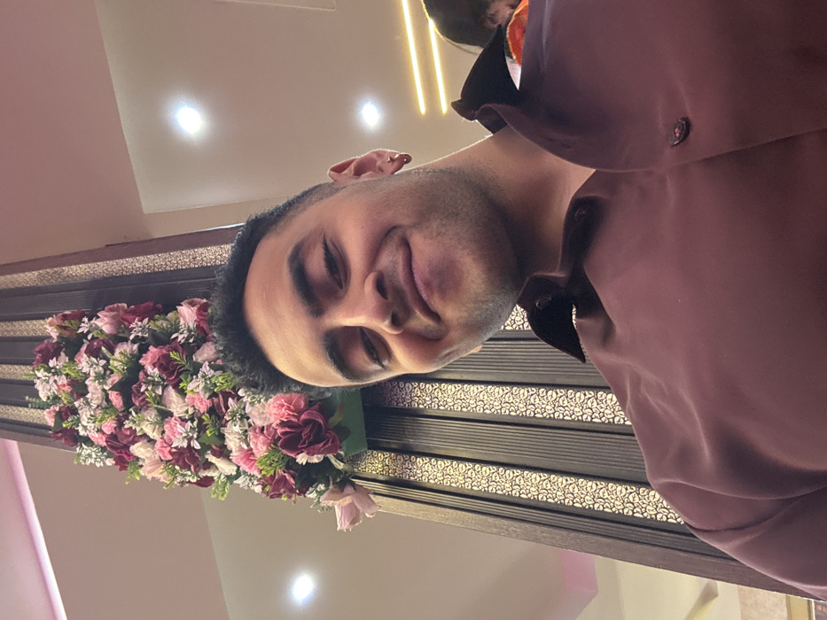 A man in a maroon collared shirt poses for a selfie. He stands in front of a decorative wall featuring horizontal metal bars with a pattern. To his left, a large flower arrangement with pink and burgundy flowers is visible. The background includes a light pink wall and a white ceiling with several recessed lights. The man has short dark hair and a slight smile.