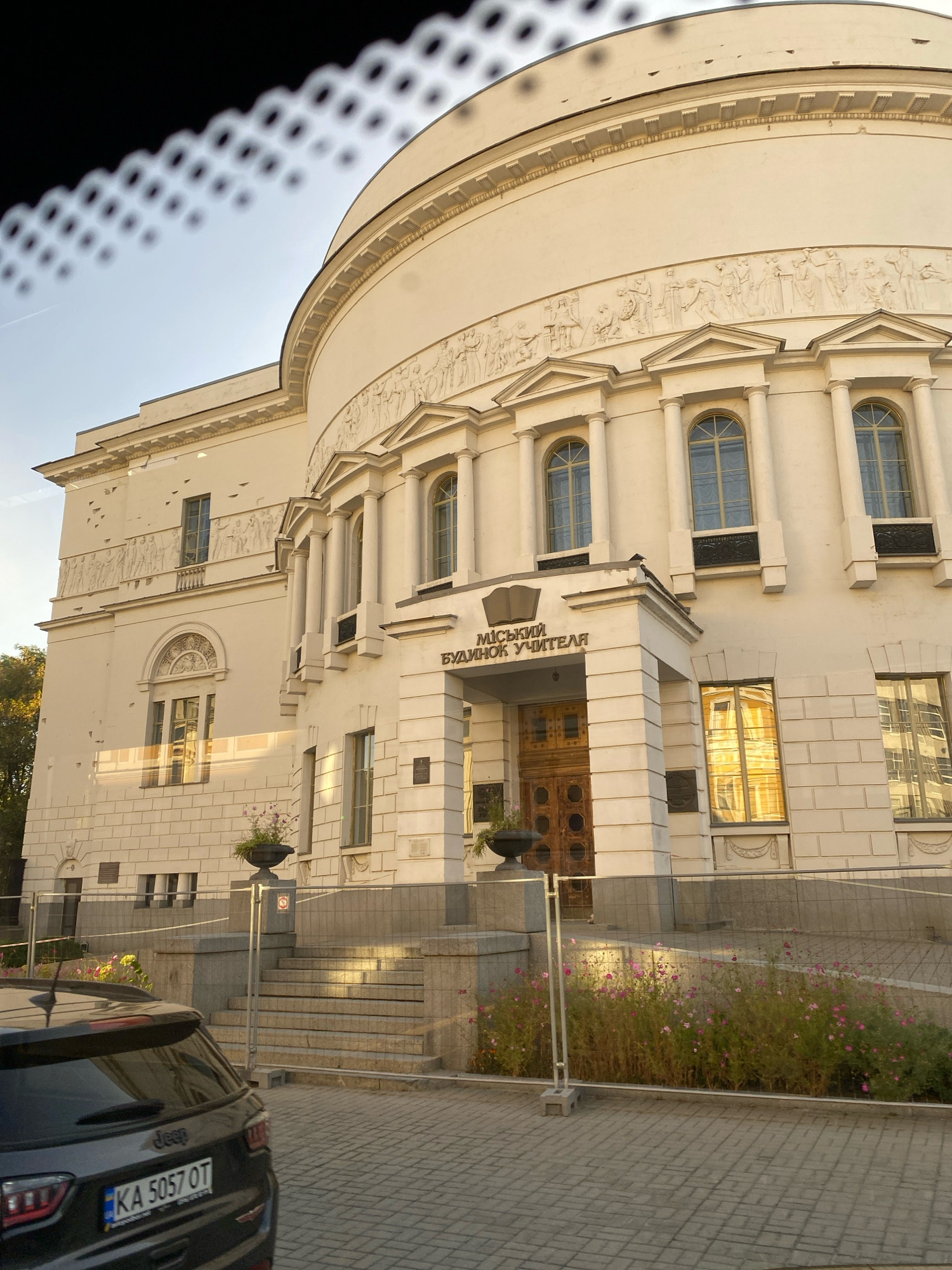 A light-colored, multi-story building with classical architectural elements is seen from a low angle, partially obscured by what appears to be a reflection or overlay on the lens. The building features a curved facade on the right, adorned with a frieze of figures in relief. Numerous arched windows with decorative grilles are visible. A central entrance is framed by columns and surmounted by a pediment with an open book emblem and text in Cyrillic, "МІСЬКИЙ БУДИНОК УЧИТЕЛЯ". To the left, a smaller section of the building has a flat facade with more windows and a semicircular decorative element above one window. In the foreground, the rear of a dark-colored SUV with a license plate reading "KA 5057 OT" is parked on a cobblestone surface. A set of stone stairs leads up to the building's entrance, bordered by low metal fencing and planters with pink flowers. The sky is a clear blue, with a faint contrail visible.