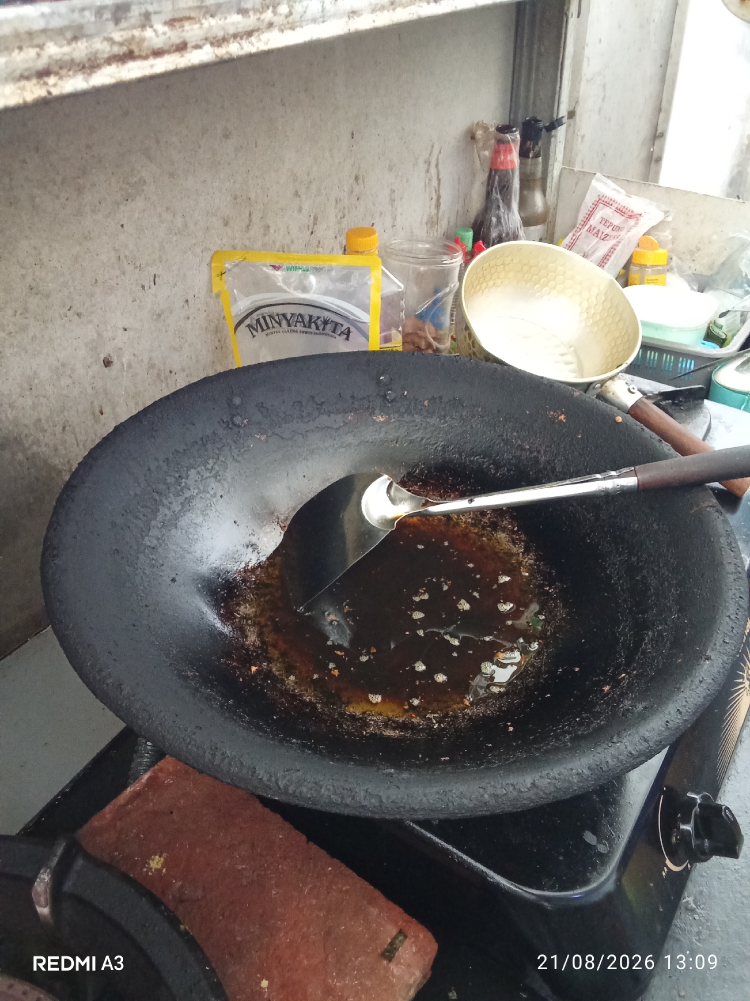 An overhead, medium shot captures a used black wok with cooking oil and food residue on a stovetop. A silver ladle with a dark wooden handle rests inside the wok. To the left, a red brick lies on the stovetop. In the background, on a shelf, are several items including a plastic bag labeled "MINYAKYTA", a yellow bottle, a glass jar, a light-colored textured bowl, and other containers. The setting appears to be an outdoor kitchen or food stall in Banaran, Indonesia, indicated by the timestamp "21/08/2026 13:09".