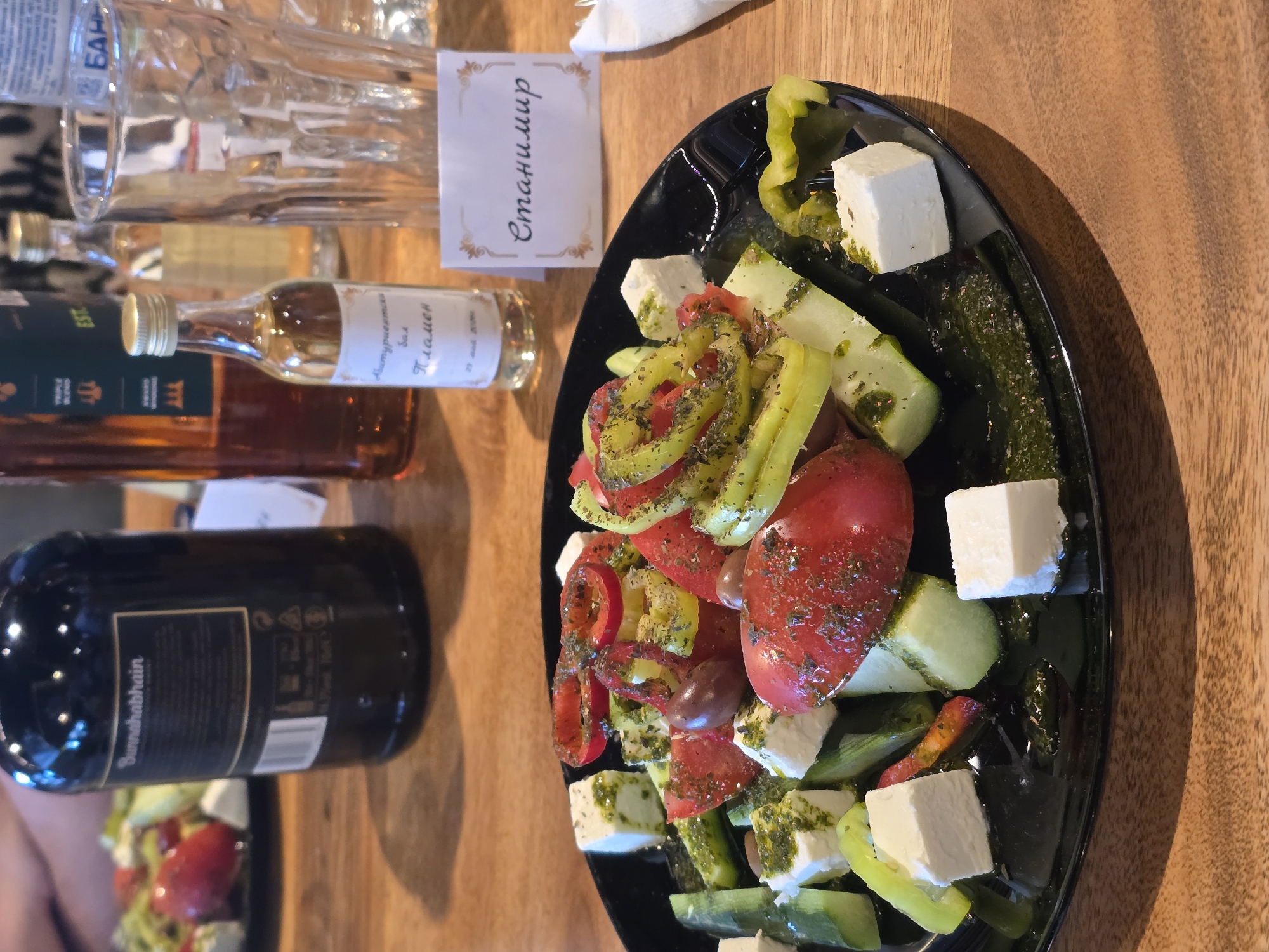 The image shows a wooden table setting with a black plate of salad, possibly Greek salad, featuring tomatoes, cucumbers, bell peppers (yellow and red), olives, and cubed feta cheese, sprinkled with herbs. A name card in Cyrillic ("Станимир") is near the plate. Behind the plate are several bottles, including a dark bottle labeled "Sunnahabhain" and two smaller bottles with light-colored liquid. Crystal glasses are also visible. The location appears to be a restaurant or a private event setup, possibly in an unidentified city.