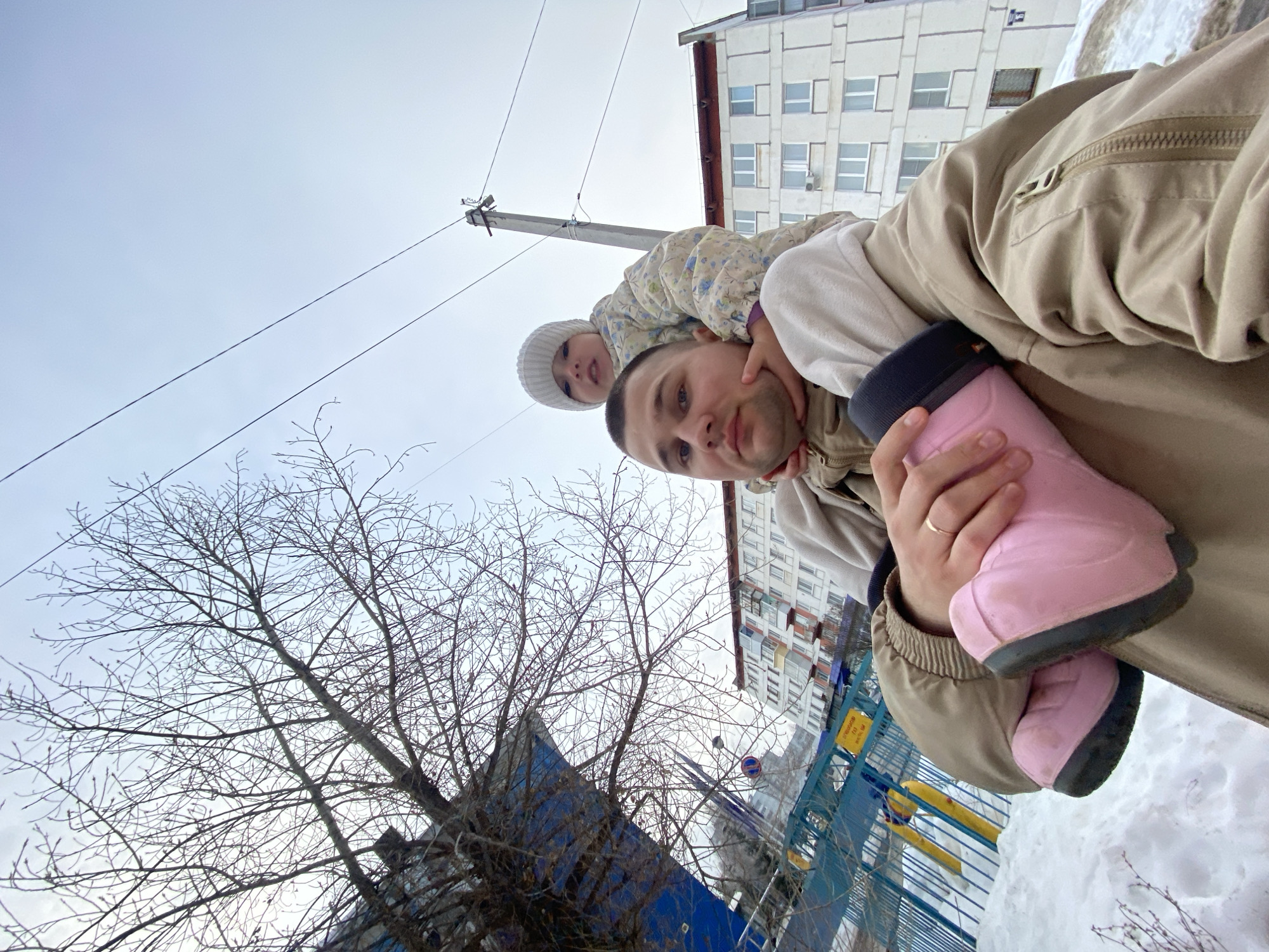 A low-angle, selfie-style photograph depicts an adult male carrying a young child on his shoulders in a snowy, overcast urban environment. The man, positioned centrally, has short hair, a clean-shaven face, and wears a light brown winter jacket over a lighter hooded garment. A gold band is visible on the ring finger of his left hand, which is holding one of the child's prominent pink boots with black soles. The child, seated on the man's shoulders, wears a white knit winter hat and a light-colored patterned puffer jacket. The child's legs are wrapped around the man's head, and hands rest on the man's face, with both subjects looking directly towards the camera.
The background reveals patches of snow covering the ground, indicating cold weather. To the left, a large deciduous tree with bare branches occupies the upper left quadrant, adjacent to a dark blue structure, possibly a building facade or a tall fence. Behind this, a blue metal fence and yellow playground equipment are partially visible. A yellow sign with Cyrillic text, including "ОСТОРОЖНО ГАЗ" (CAUTION GAS), and a circular traffic sign are partially visible through the fence. In the mid-ground, several multi-story residential apartment buildings are present, primarily white with multiple windows. On the right side, a grey concrete utility pole with power lines extends vertically against a pale, overcast sky. The scene is located in a residential area of Chaikovsky, Russia.
