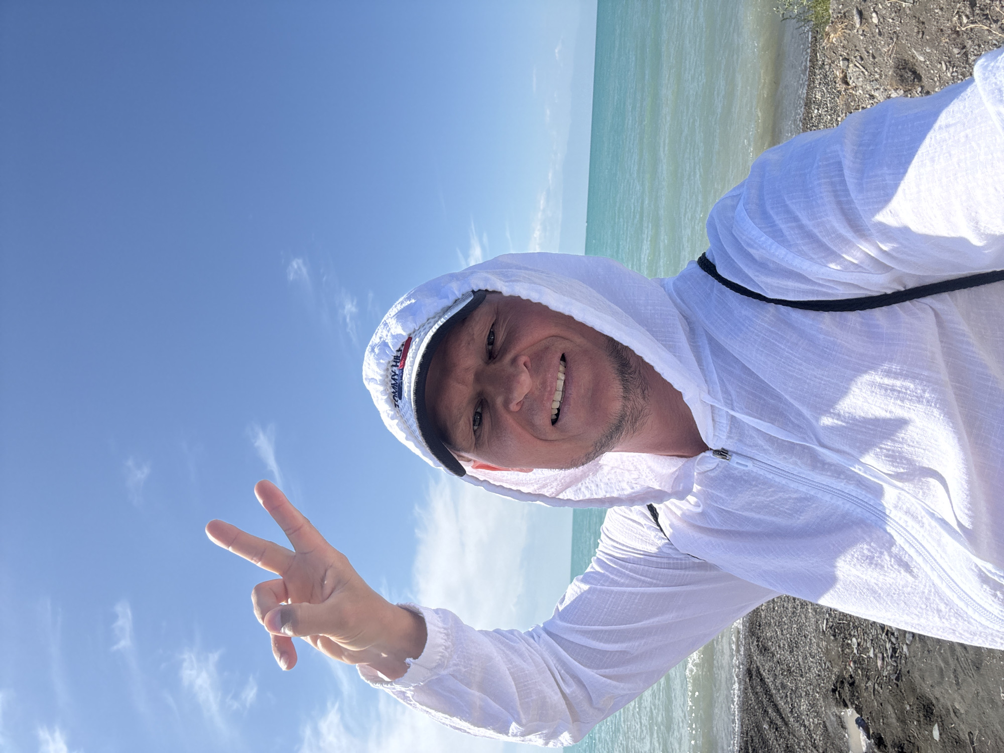 A man, wearing a white hooded jacket and a white baseball cap, stands on a rocky shore, holding up his left hand in a peace sign. He is smiling at the camera, and his face is lit by sunlight. The sea, with its blue-green water, stretches out behind him under a bright blue sky with scattered white clouds. The shoreline is composed of dark pebbles and sand. The man appears to be in Zharbulak, Kazakhstan.